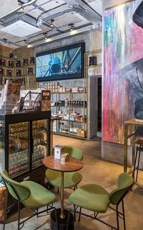 Interno di un moderno bar con tavolo rotondo e quattro sedie verdi, grande quadro colorato sulla parete e scaffali con prodotti e bottiglie.