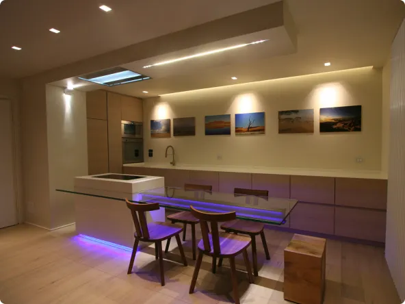 Cucina moderna con isola centrale, tavolo con piano in vetro e sedie in legno, illuminazione soffusa e decorazioni fotografiche sulle pareti.