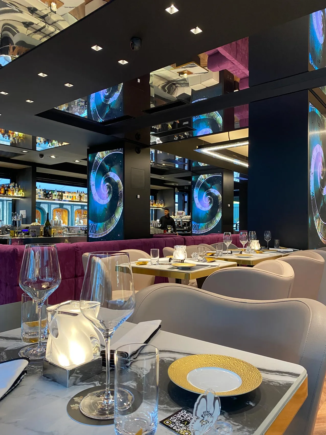 Interno elegante di un ristorante con divano viola in primo piano, tavoli rotondi apparecchiati e illuminazione moderna.