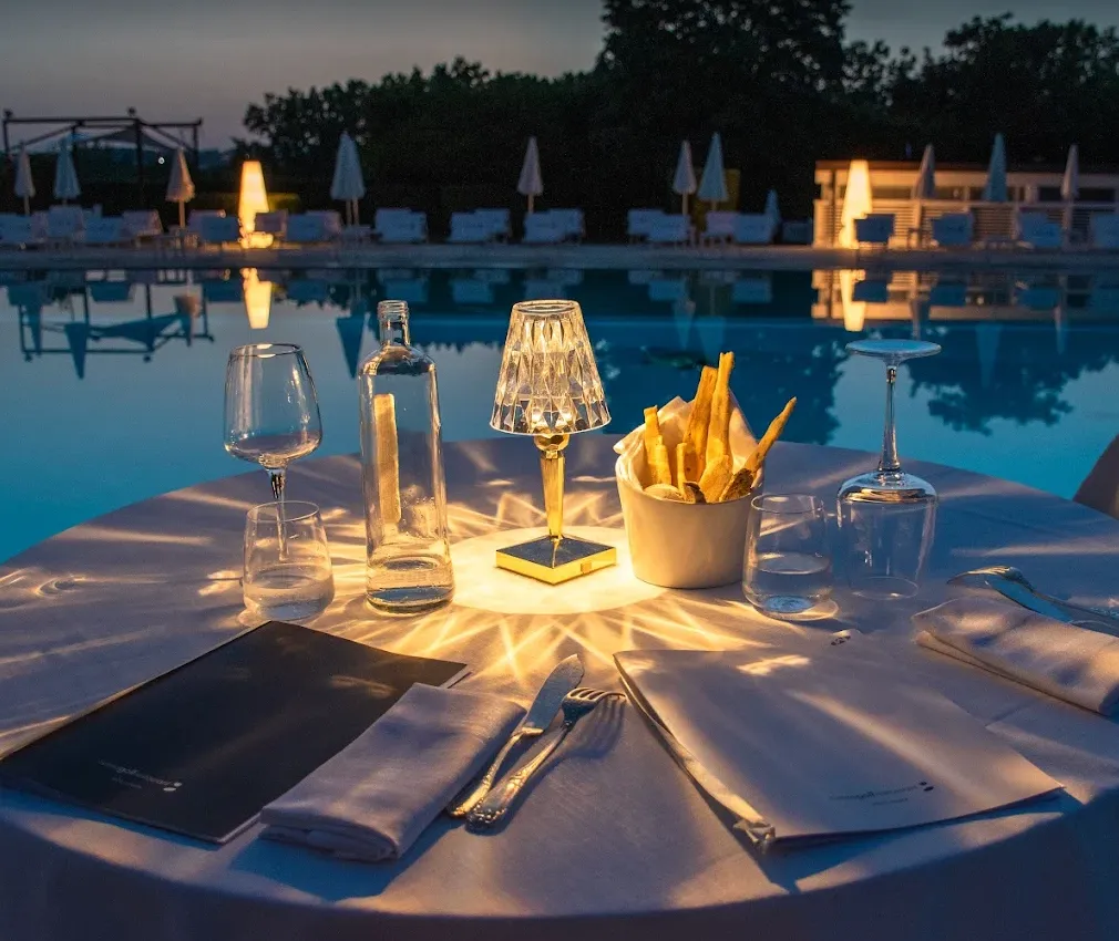 Tavolo da cena all'aperto illuminato da una lampada con vista su una piscina di notte, con un bicchiere d'acqua, una bottiglia, un contenitore con patatine e stoviglie.