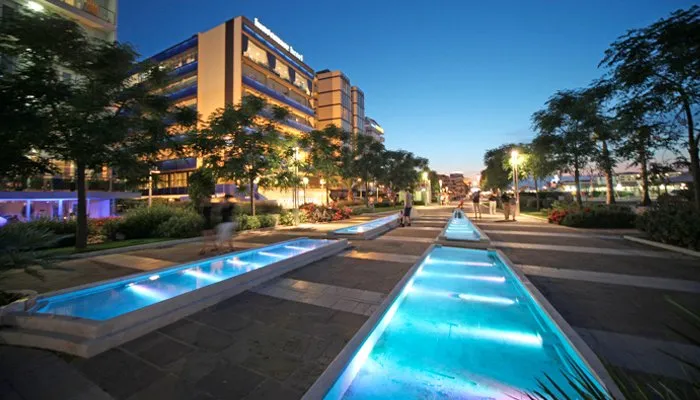 Viale serale illuminato con fontane d'acqua blu e alberi, con edifici moderni sullo sfondo.