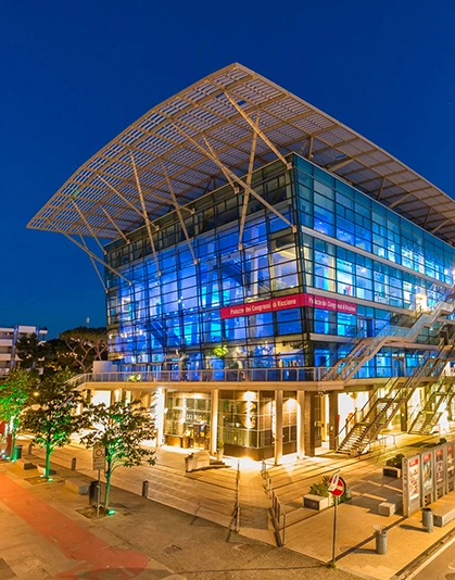 Edificio moderno con facciata in vetro illuminato di blu la sera, con tetto a griglia e scala esterna.