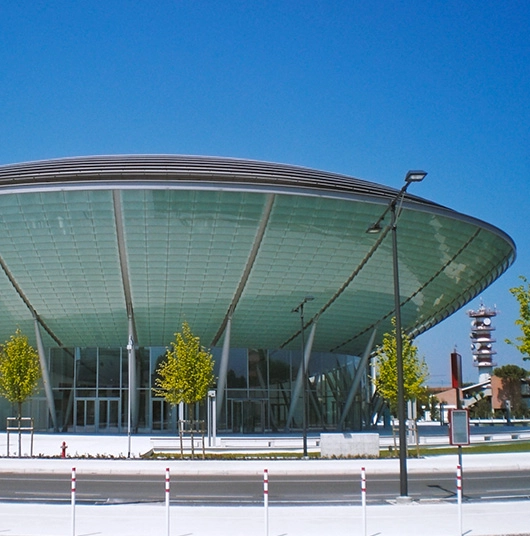 Edificio moderno con tetto a forma di disco e grandi vetrate, circondato da alberi e lampioni sotto un cielo azzurro.