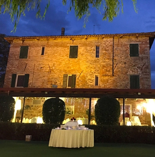 Antica casa in pietra illuminata di sera con un tavolo rotondo apparecchiato in giardino davanti.