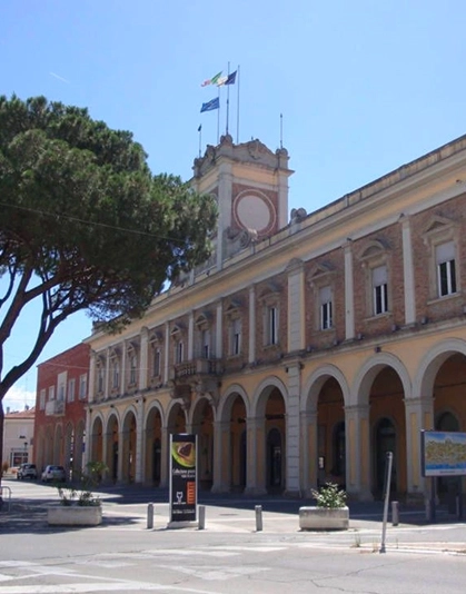 Edificio storico con una torre centrale con tre bandiere e una fila di archi sul piano terra, con alberi e cielo sereno.