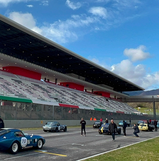 Auto d'epoca parcheggiate su una pista di gara davanti a tribune vuote di uno stadio sotto un cielo parzialmente nuvoloso.
