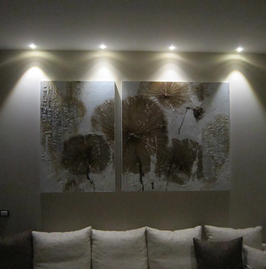 Due quadri astratti con motivi floreali marroni illuminati da quattro faretti sopra un divano chiaro con cuscini.
