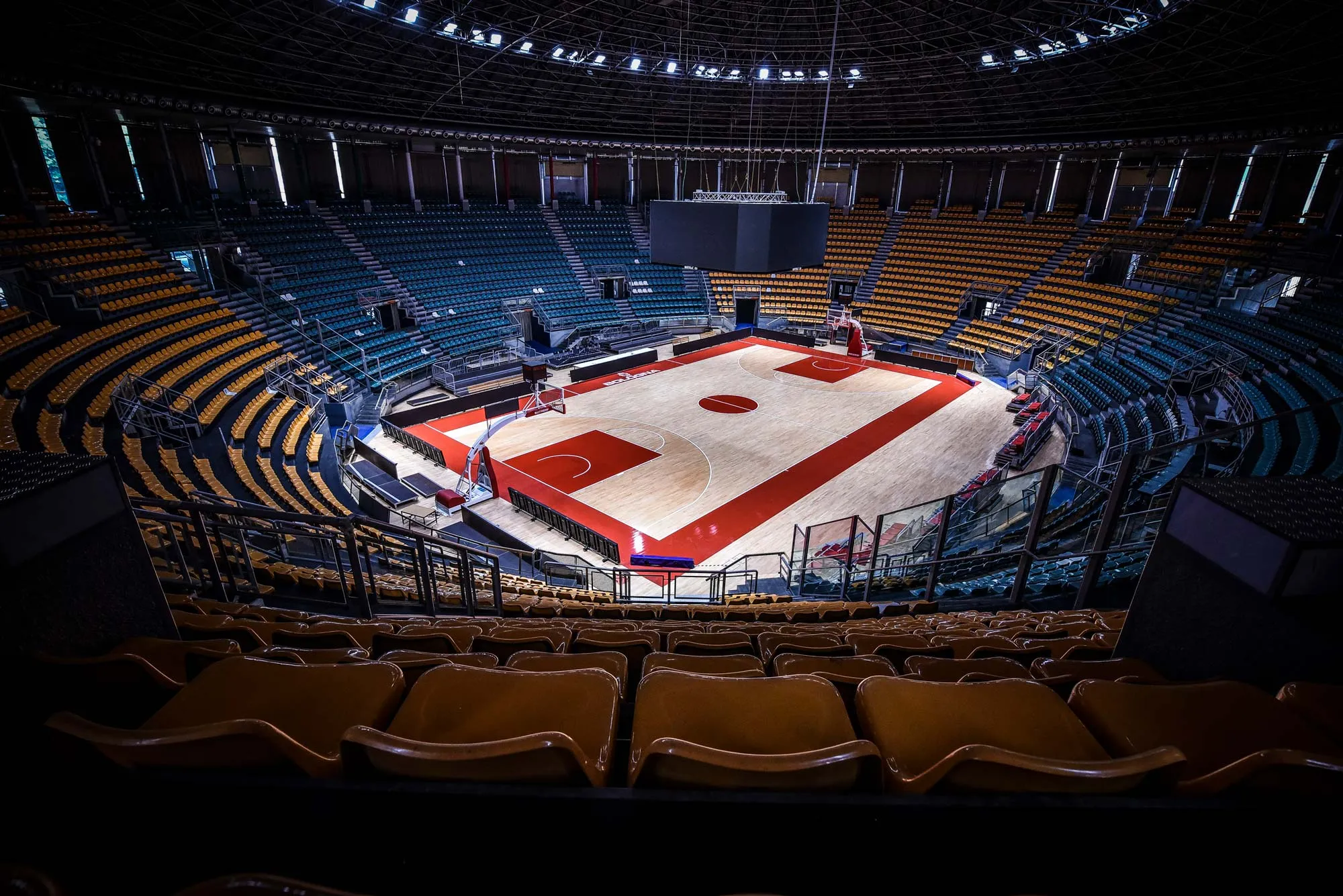 Interno di un grande palazzetto dello sport con campo da basket vuoto e gradinate con sedili gialli e verdi.