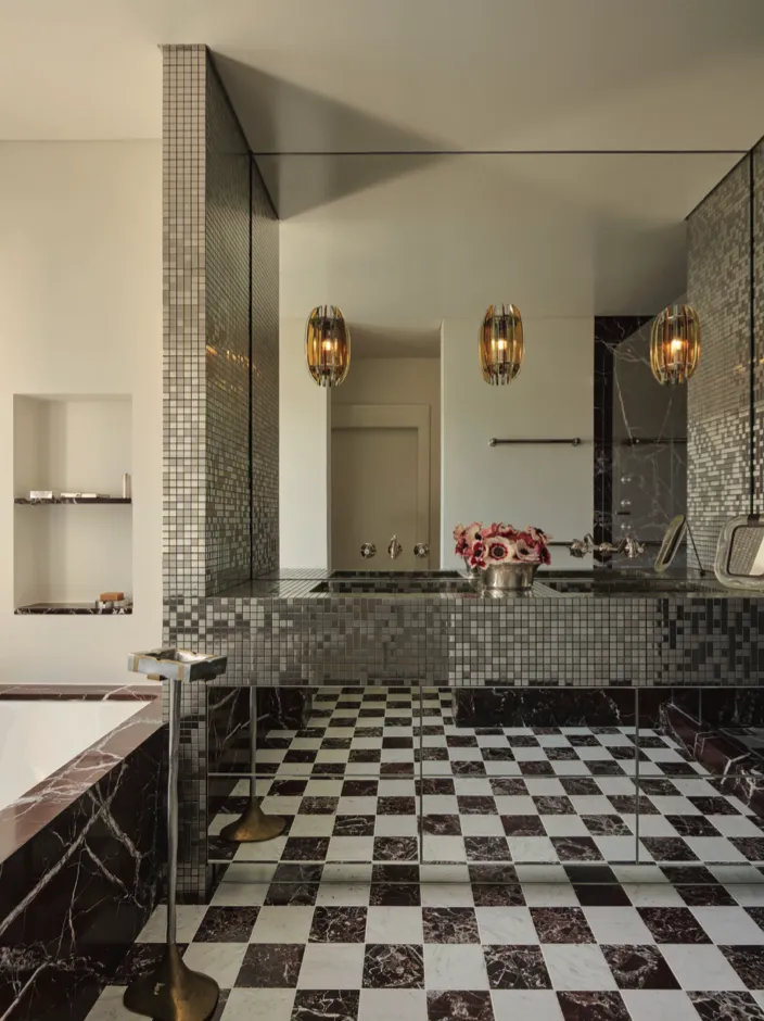 Bagno elegante con pavimento a scacchiera nero e bianco, pareti con piastrelle a mosaico e luci sospese dorate.