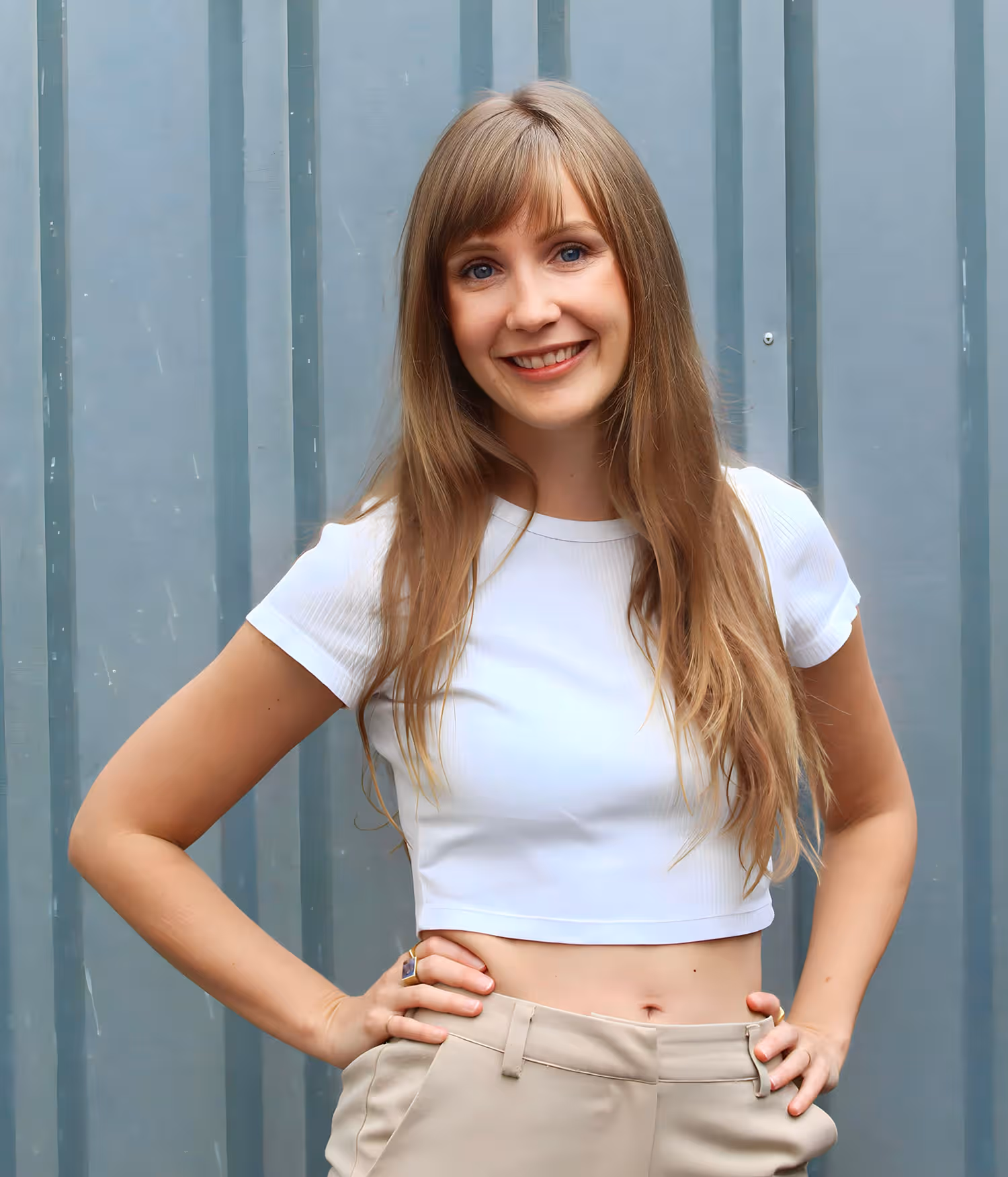 Viola smiling with long hair wearing a white crop top and beige pants standing with hands on hips.