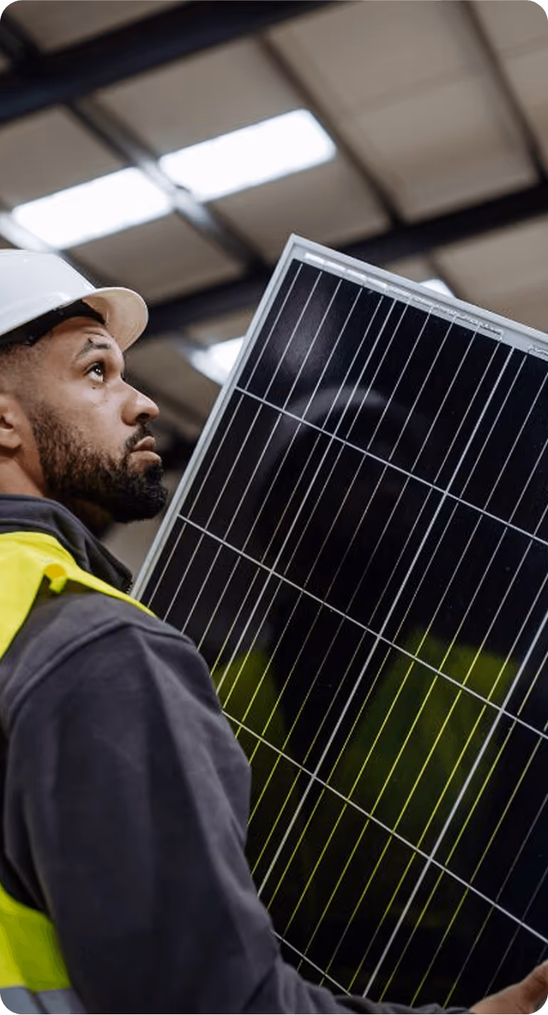 Un ouvrier portant un casque et un gilet de sécurité tient un panneau solaire dans un bâtiment.
