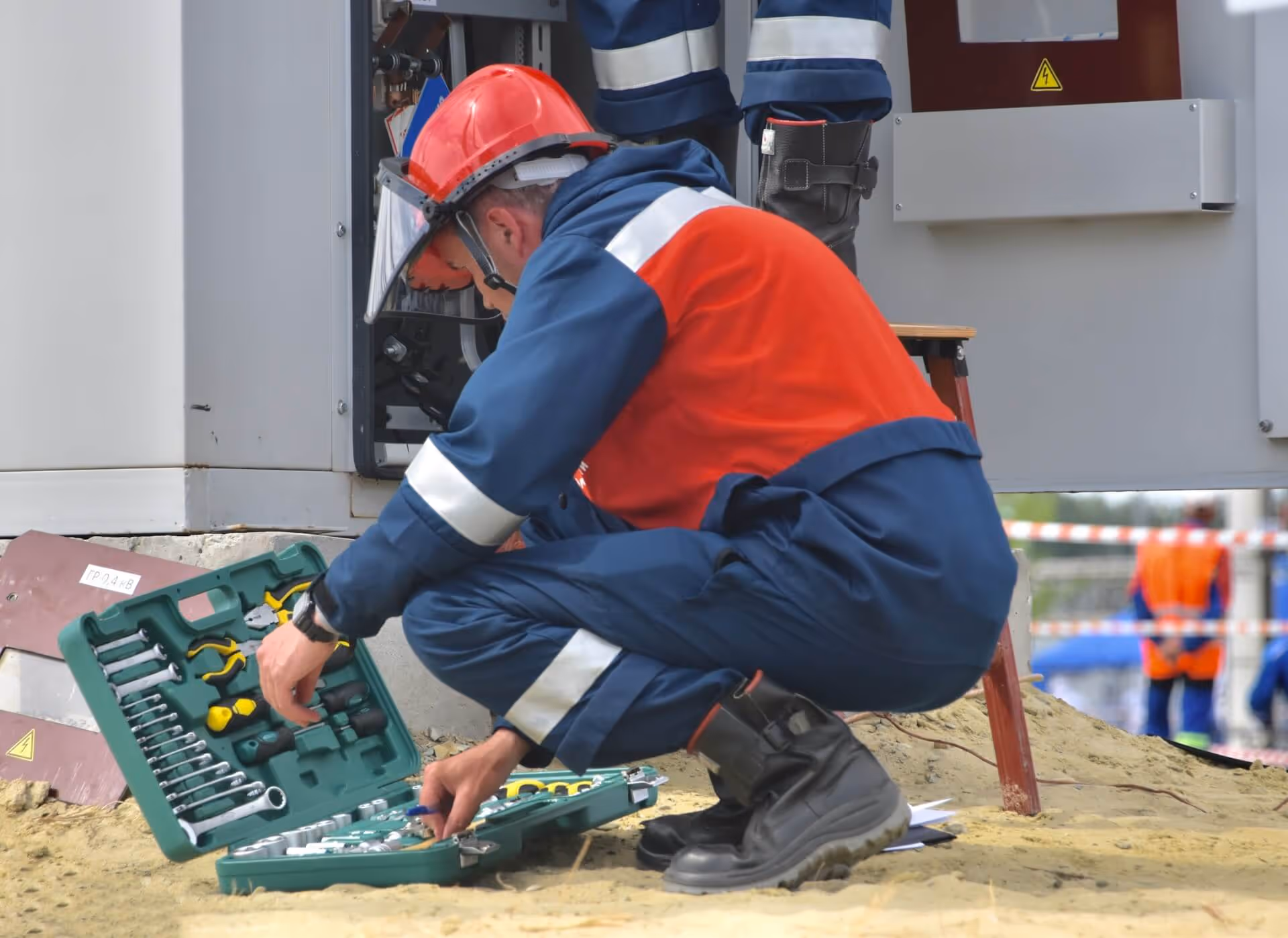 Électricien portant un casque et un équipement de sécurité, accroupi et sélectionnant un outil dans une boîte à outils verte sur un chantier extérieur.