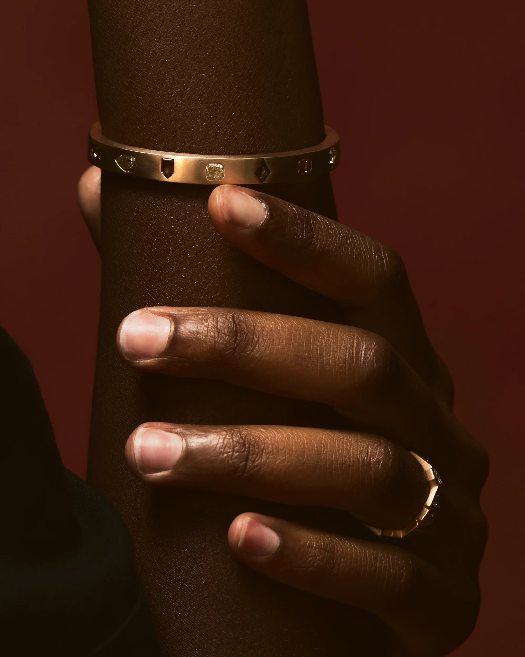 Hand wearing a gold bracelet and ring on a dark background.