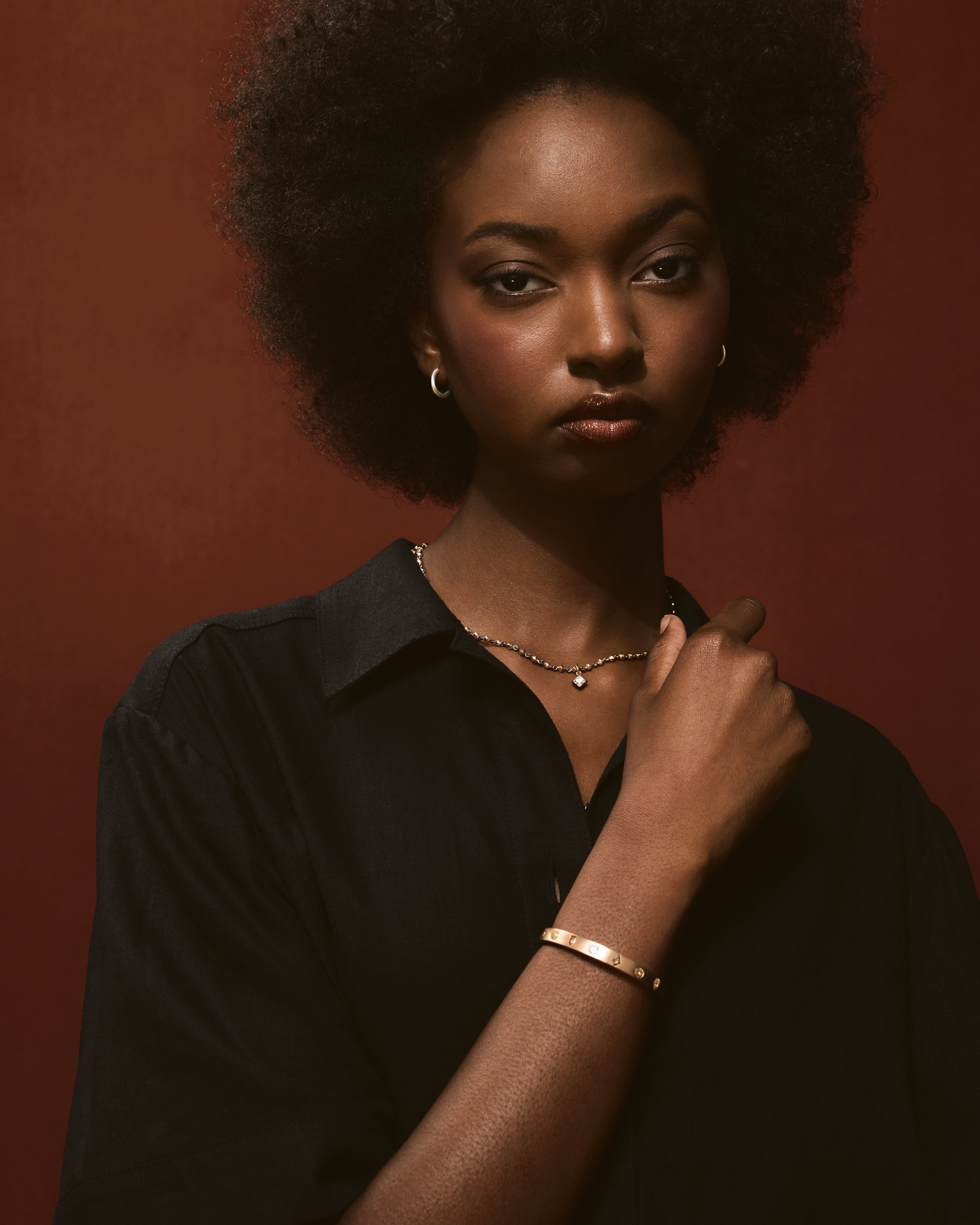 Model wearing a black shirt paired with an 18k rose gold bead necklace featuring a small diamond pendant and a matching gold bangle bracelet against a dark red background.