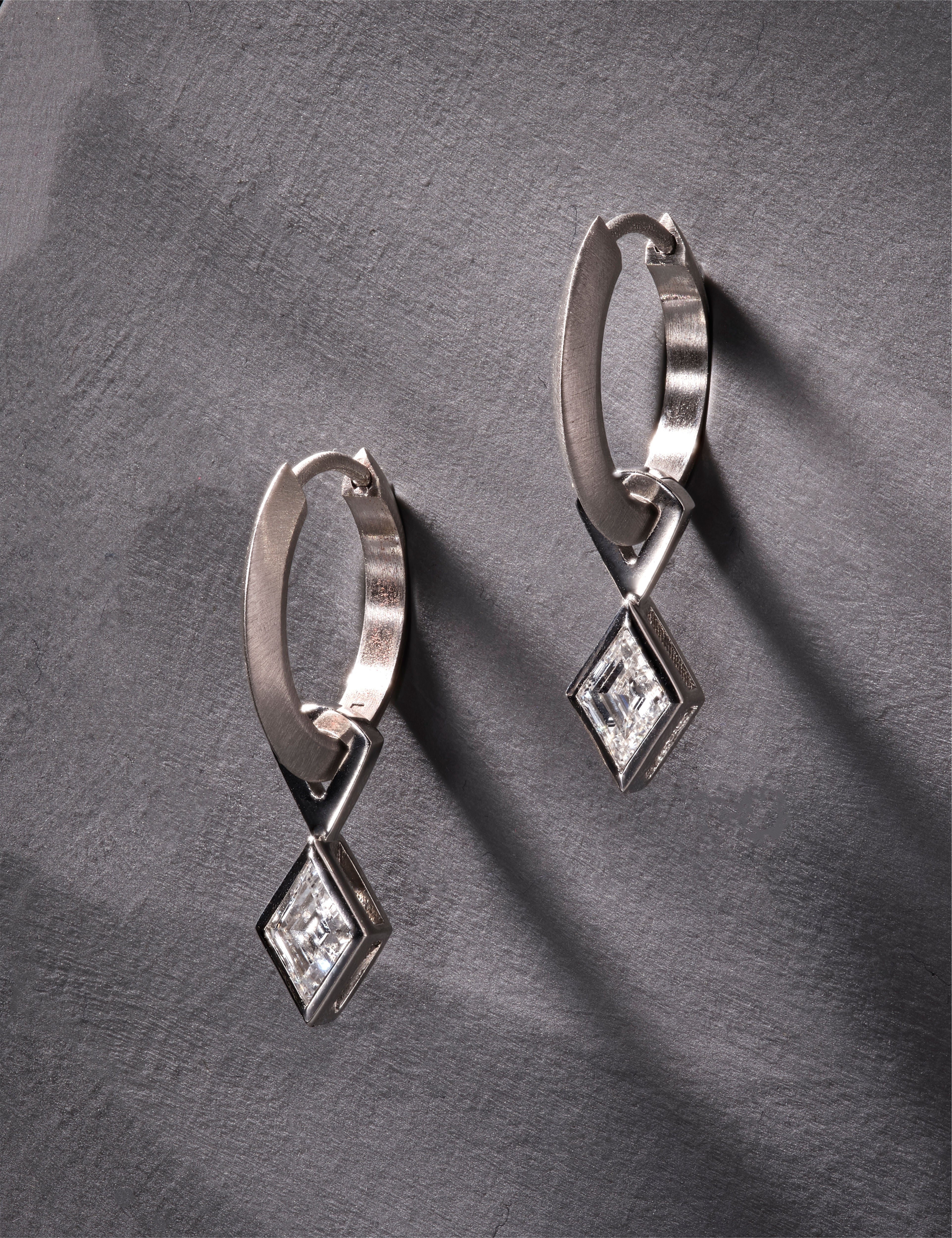 Platinum hoop earrings featuring lozenge-cut diamonds on grey textured surface.