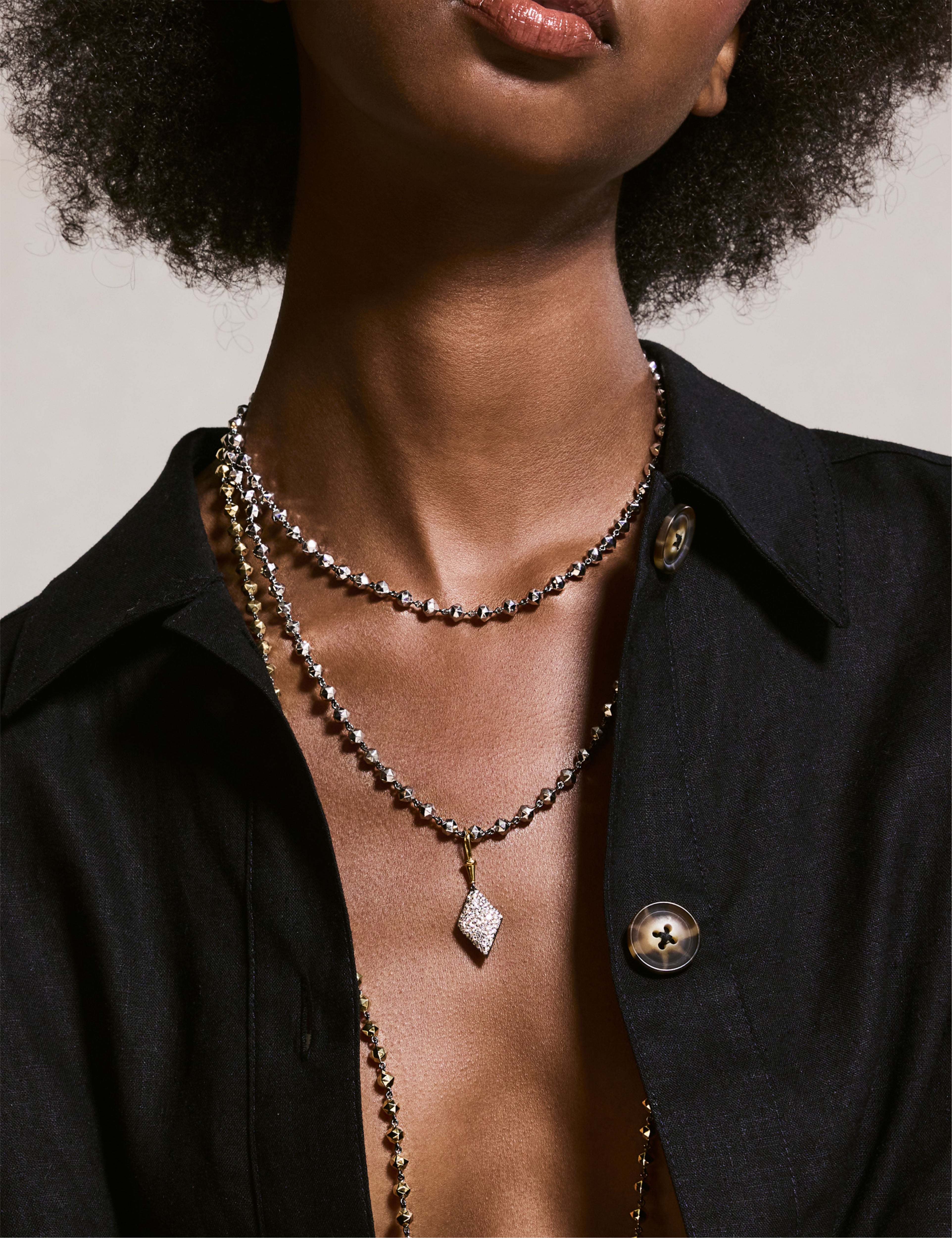 Model wearing layered gold bead necklaces with a diamond-shaped pendant and matching gold earrings, paired with a brown top.
