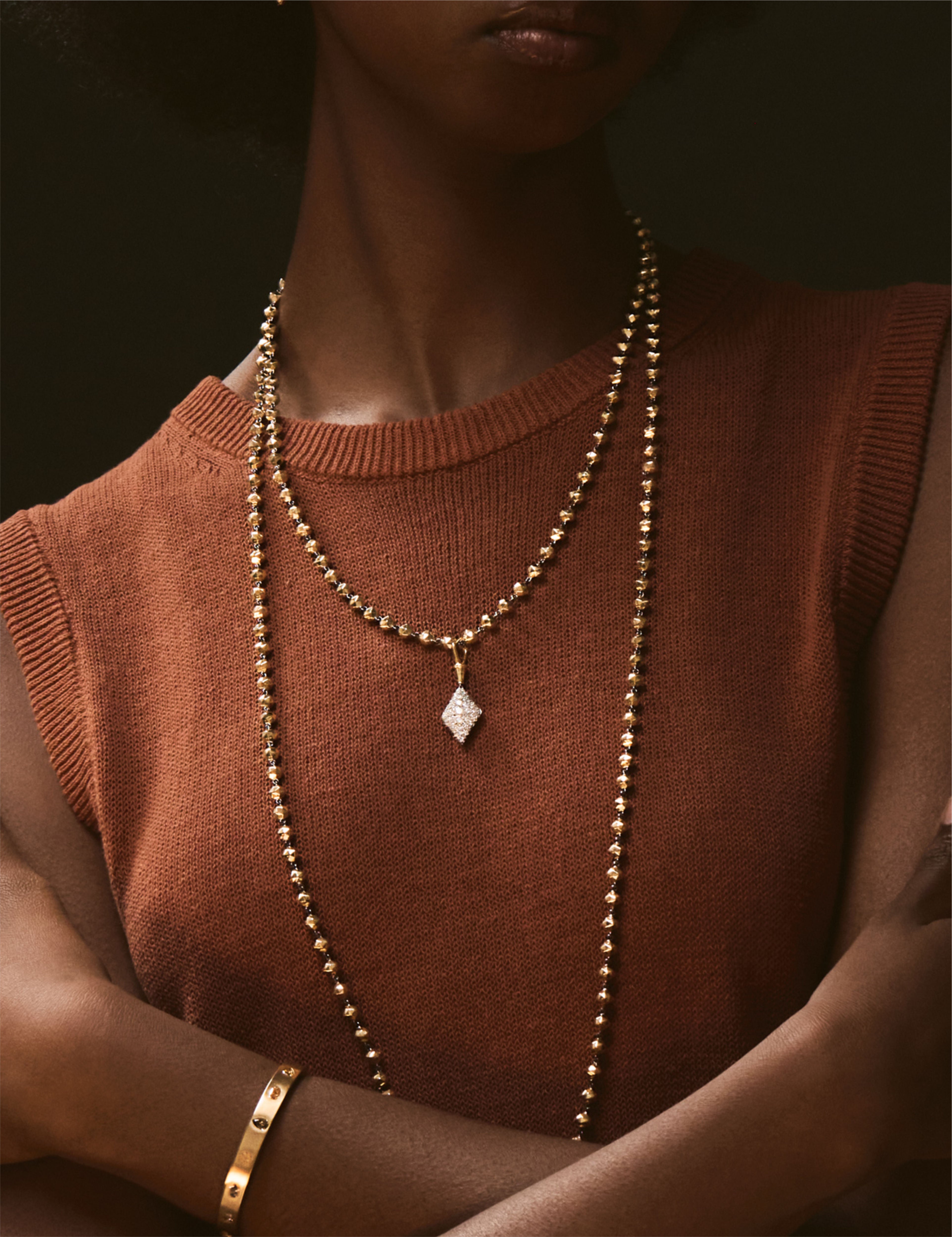 Model wearing layered gold bead necklaces with a diamond-shaped pendant and matching gold earrings, paired with a brown top.
