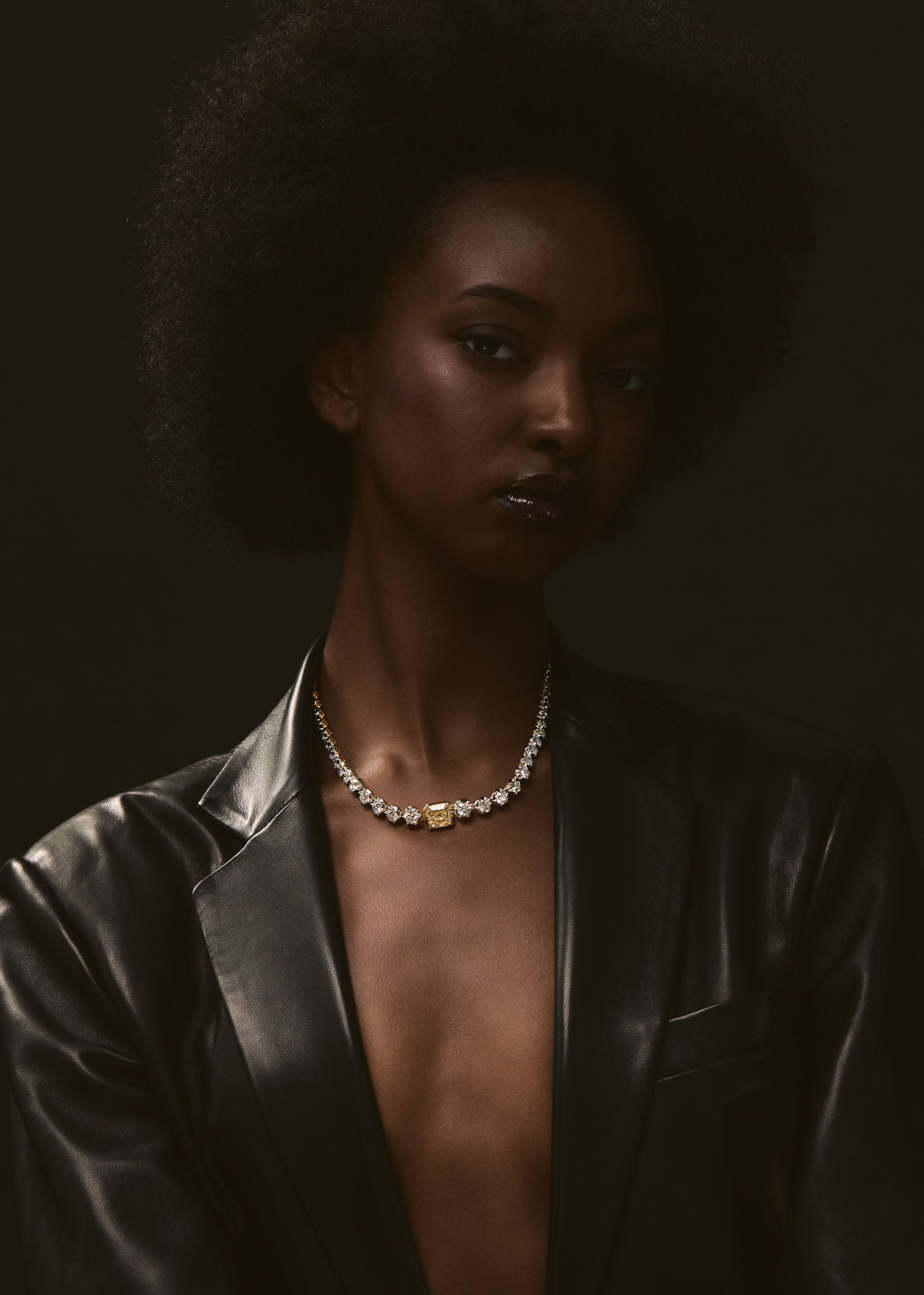 Model wearing a gold diamond necklace with a yellow diamond centerpiece styled with a black leather jacket against a dark background.