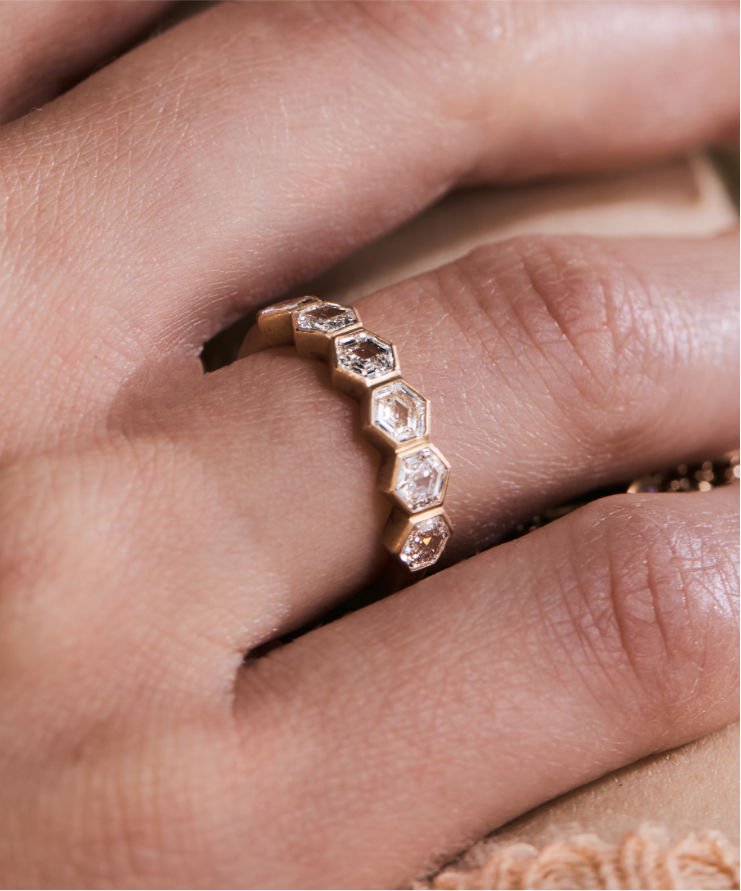 Model wearing a gold hexagon diamond eternity ring, touching hair, with bracelet detail visible in the background.