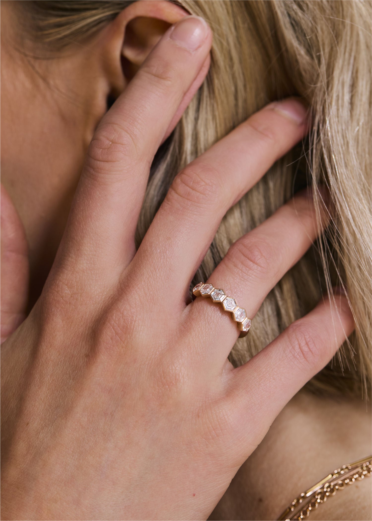 Model wearing a gold hexagon diamond eternity ring, touching hair, with bracelet detail visible in the background.