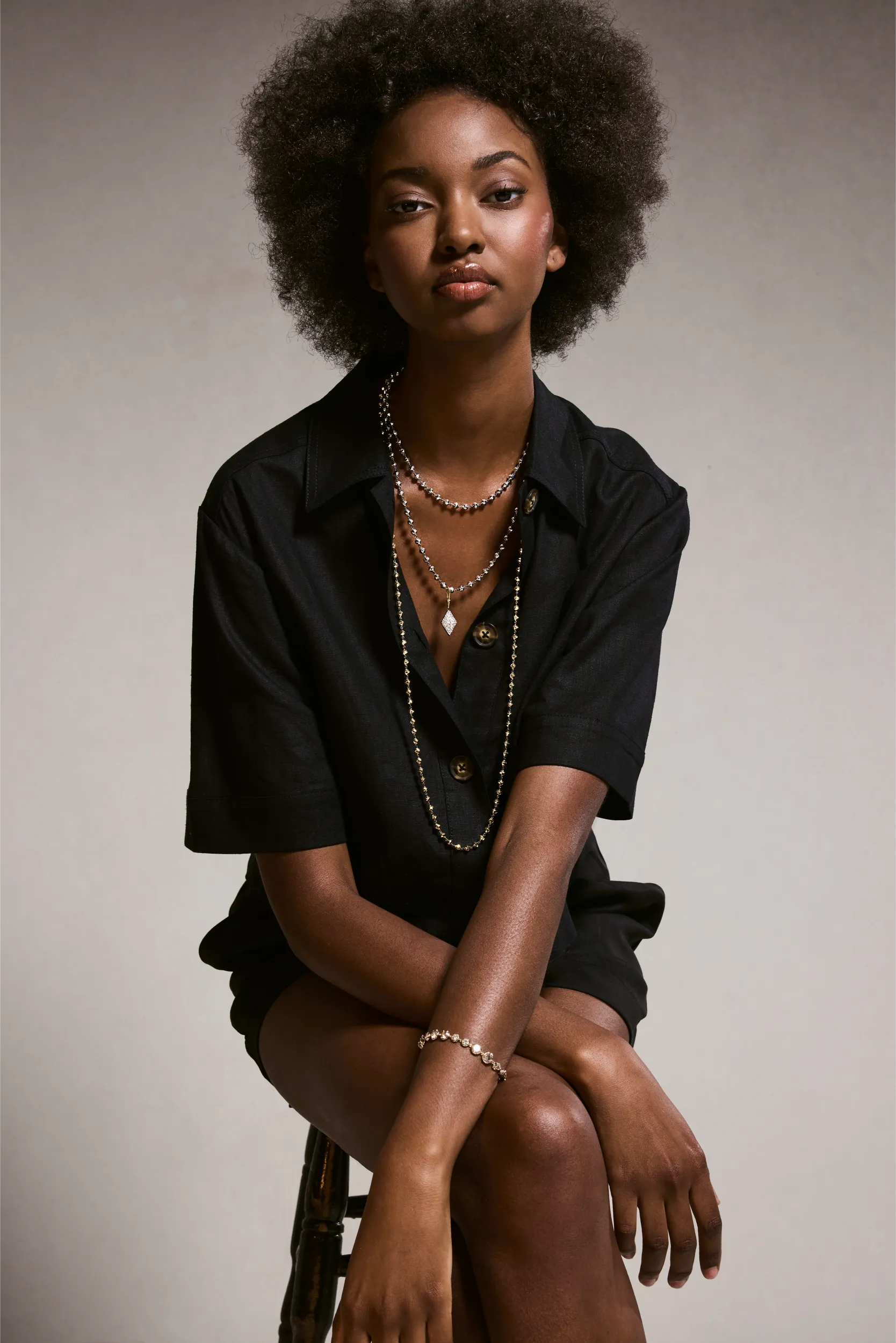 Stylish woman wearing a black outfit and layered necklaces sitting on a stool.