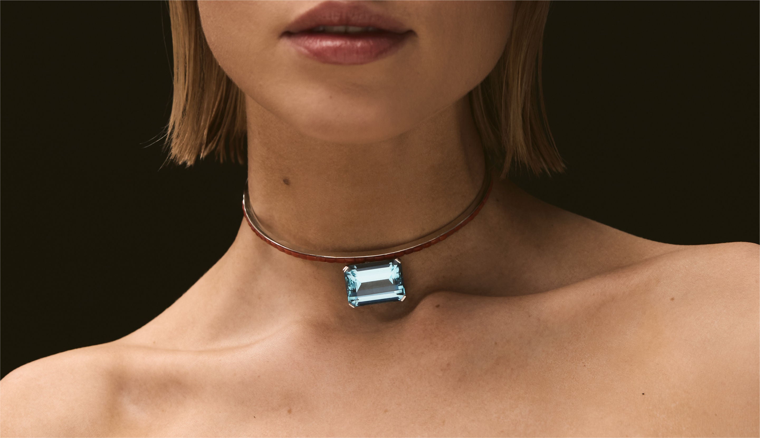 Blonde woman with slicked-back hair wearing a black strapless top and a choker necklace with a large light blue gemstone.