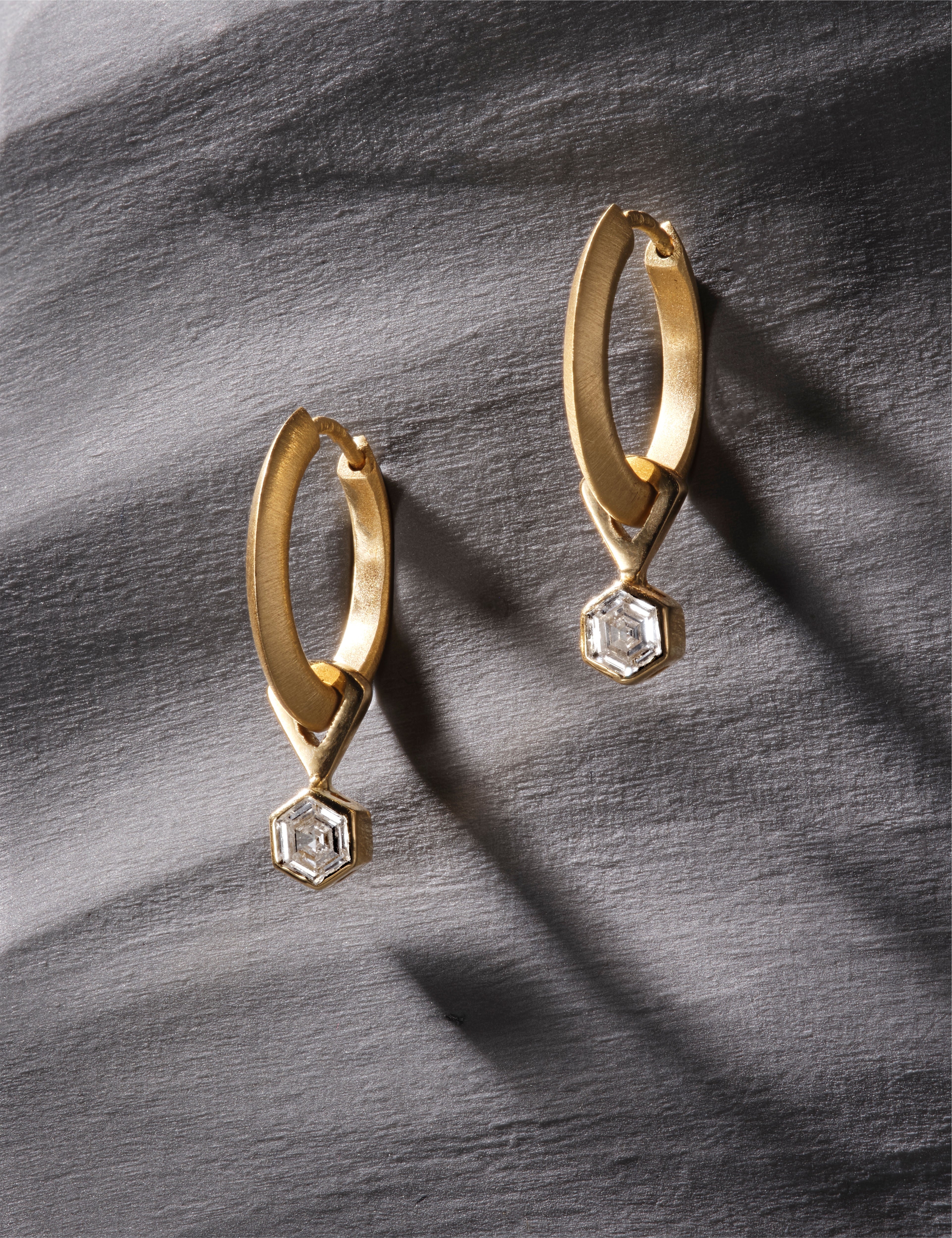 Yellow gold hoop earrings featuring hexagon-cut diamonds on grey textured surface.