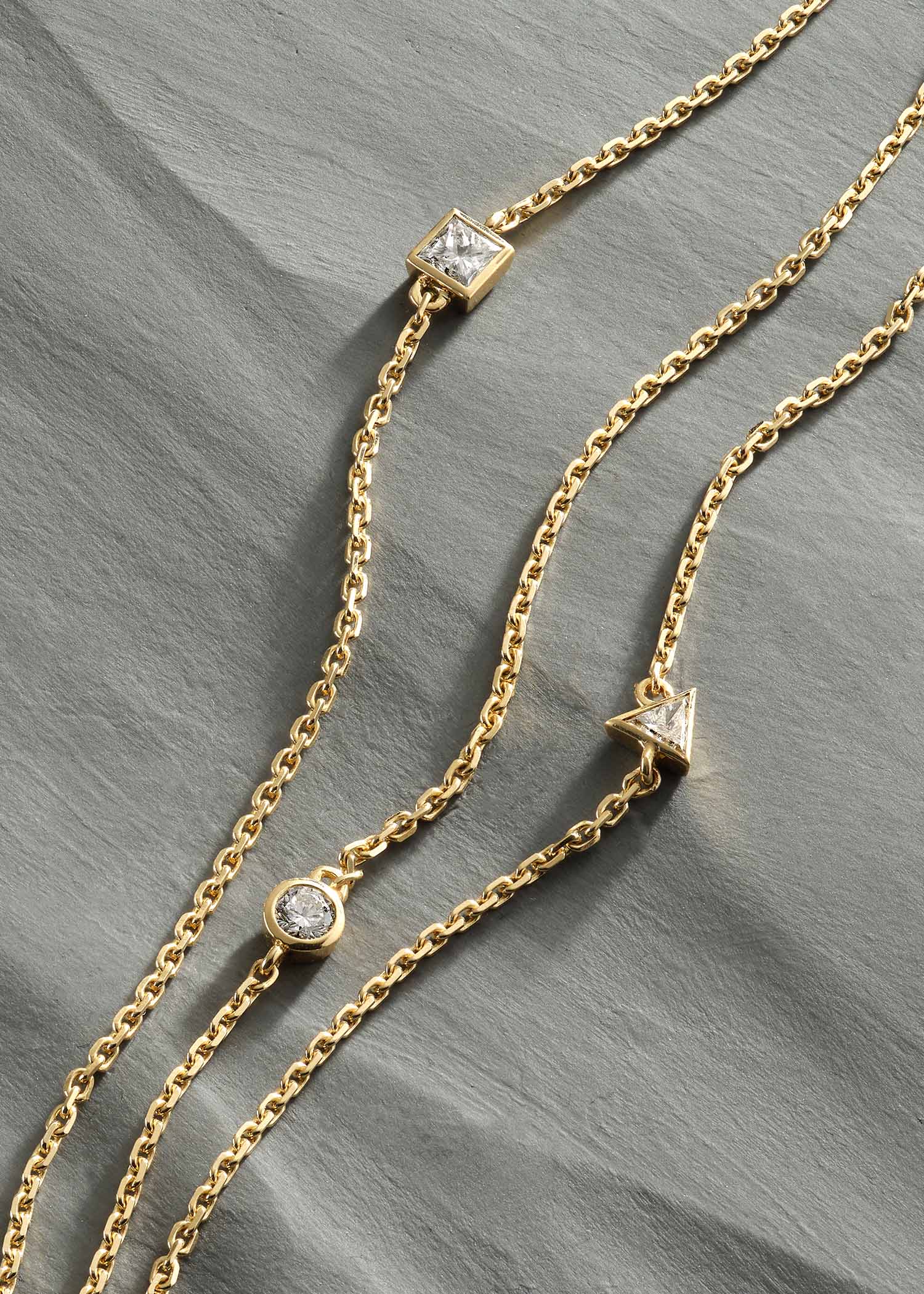 Delicate gold three-strand bracelet featuring round, triangle, and square-cut diamonds, photographed on a textured gray surface.