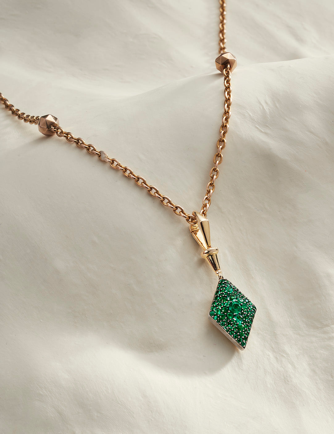 Three gold diamond-shaped pendants in green, pink, and blue gemstone settings displayed on a soft white fabric background.