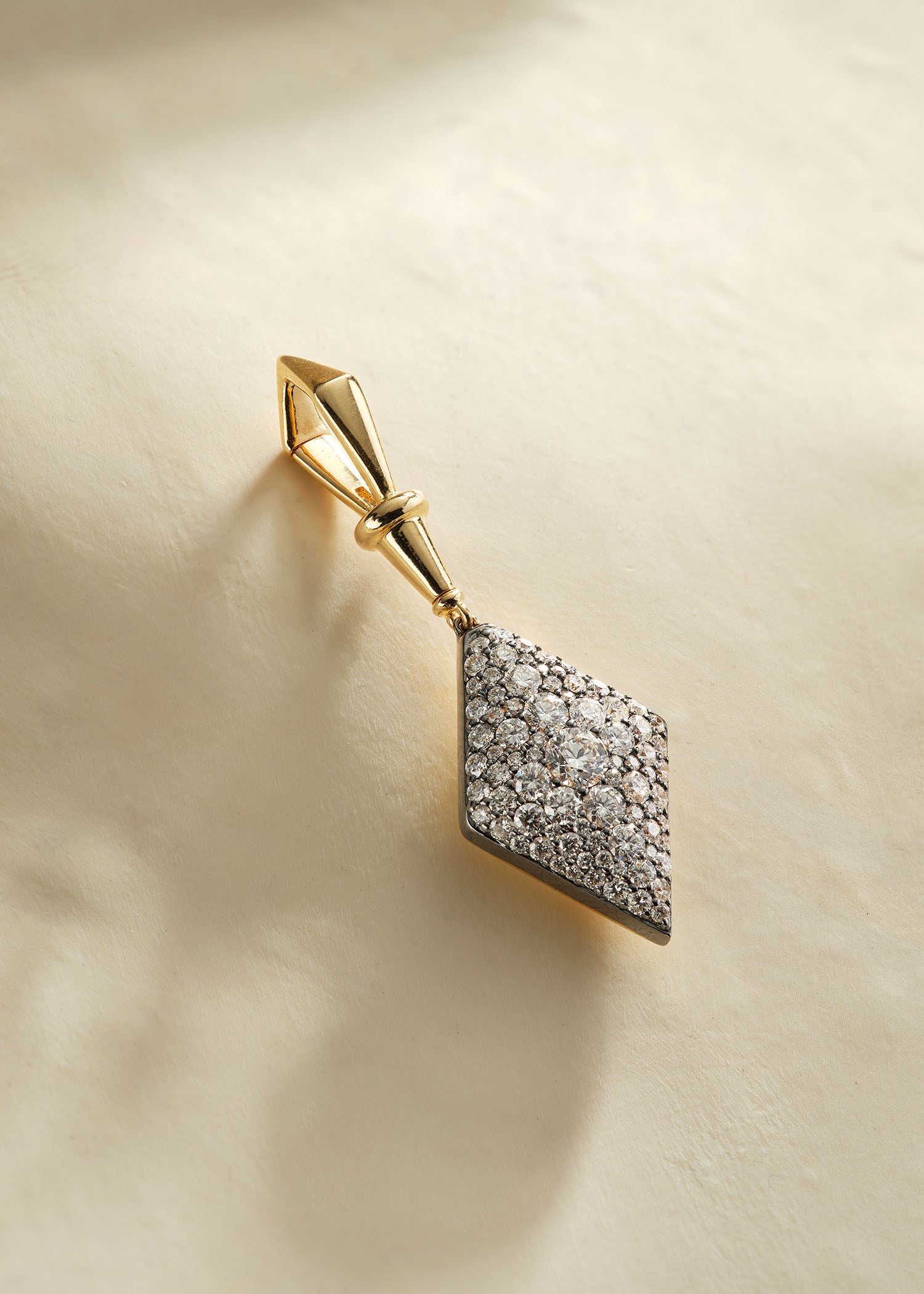 Three gold diamond-shaped pendants in green, pink, and blue gemstone settings displayed on a soft white fabric background.