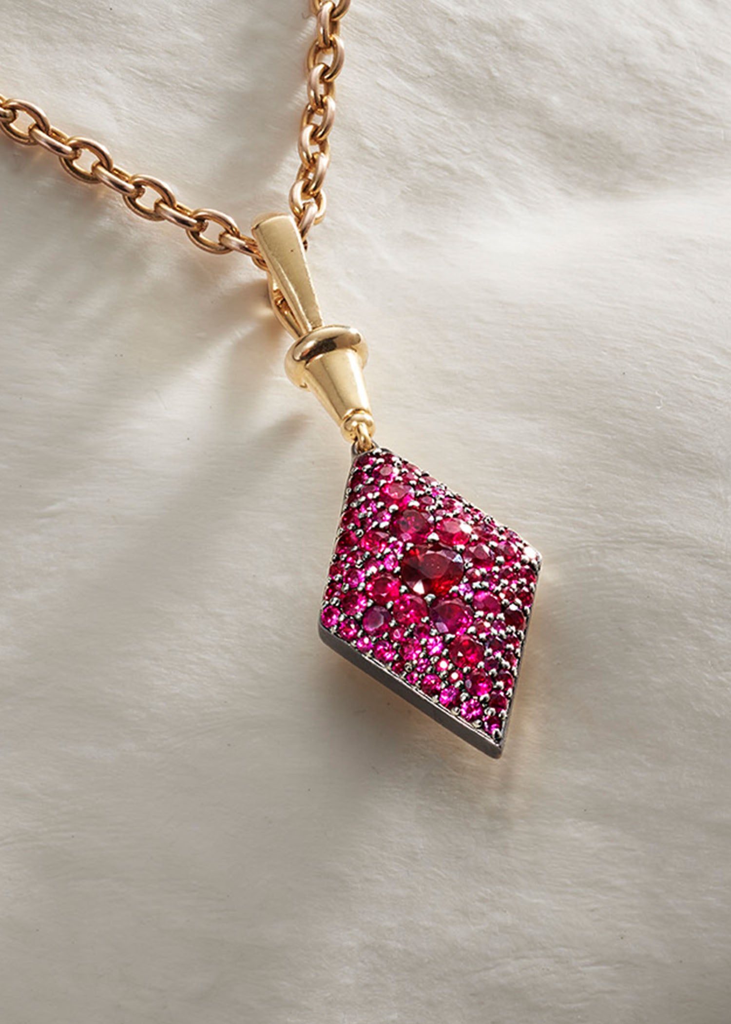 Three gold diamond-shaped pendants in green, pink, and blue gemstone settings displayed on a soft white fabric background.