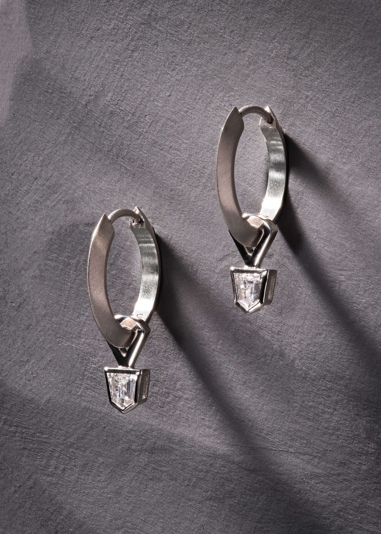 Platinum hoop earrings with geometric bullet-cut diamonds on grey textured background.