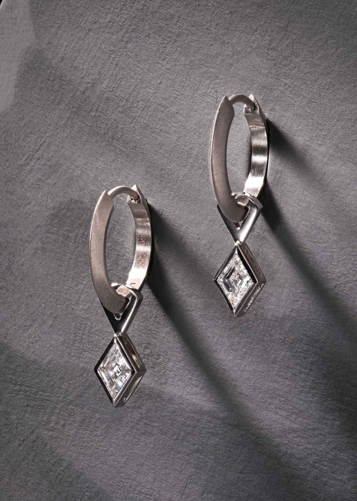 Platinum hoop earrings featuring lozenge-cut diamonds on grey textured surface.