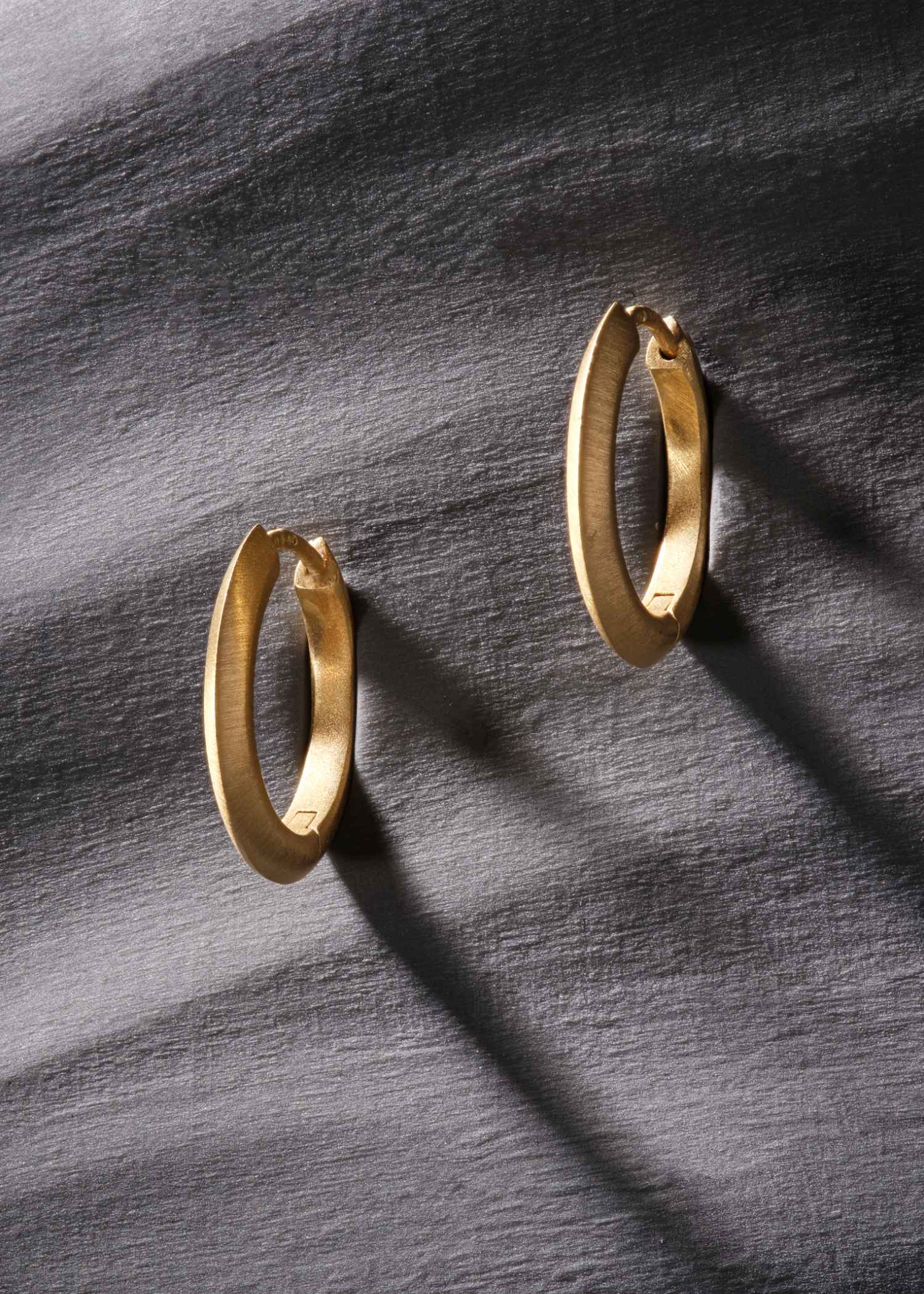 Brushed 18k yellow gold oval hoop earrings on textured grey background.