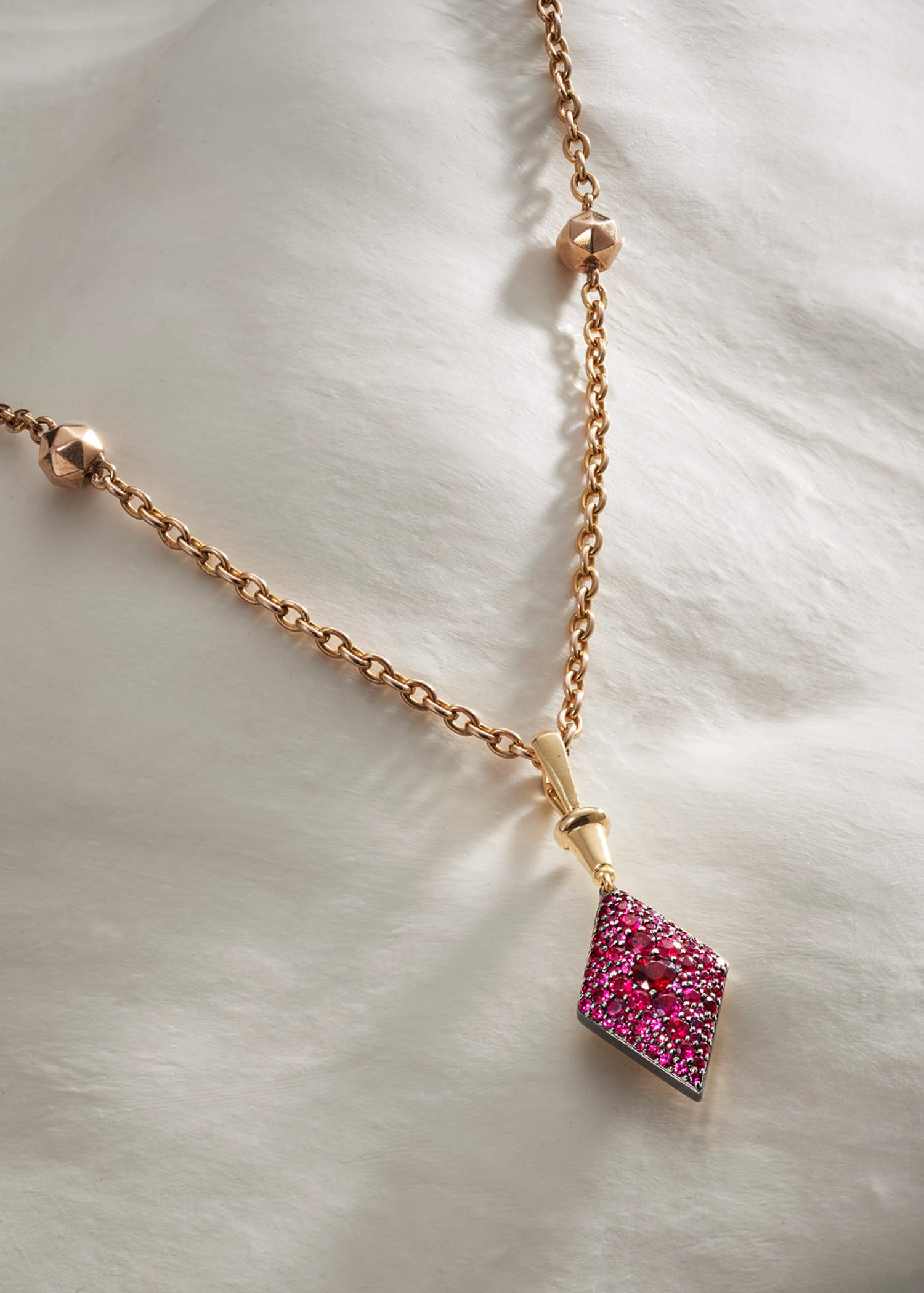 Three gold diamond-shaped pendants in green, pink, and blue gemstone settings displayed on a soft white fabric background.