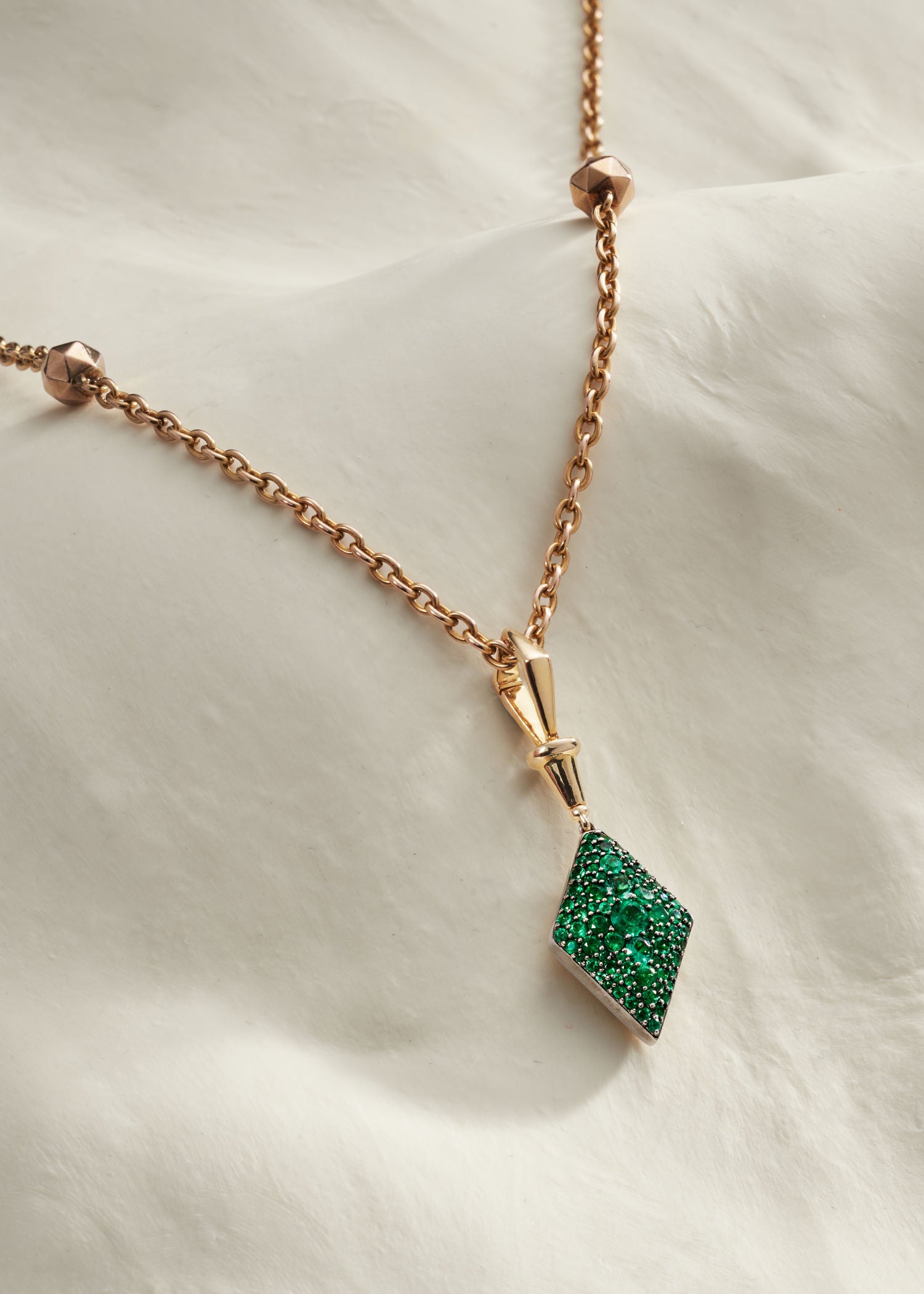 Three gold diamond-shaped pendants in green, pink, and blue gemstone settings displayed on a soft white fabric background.