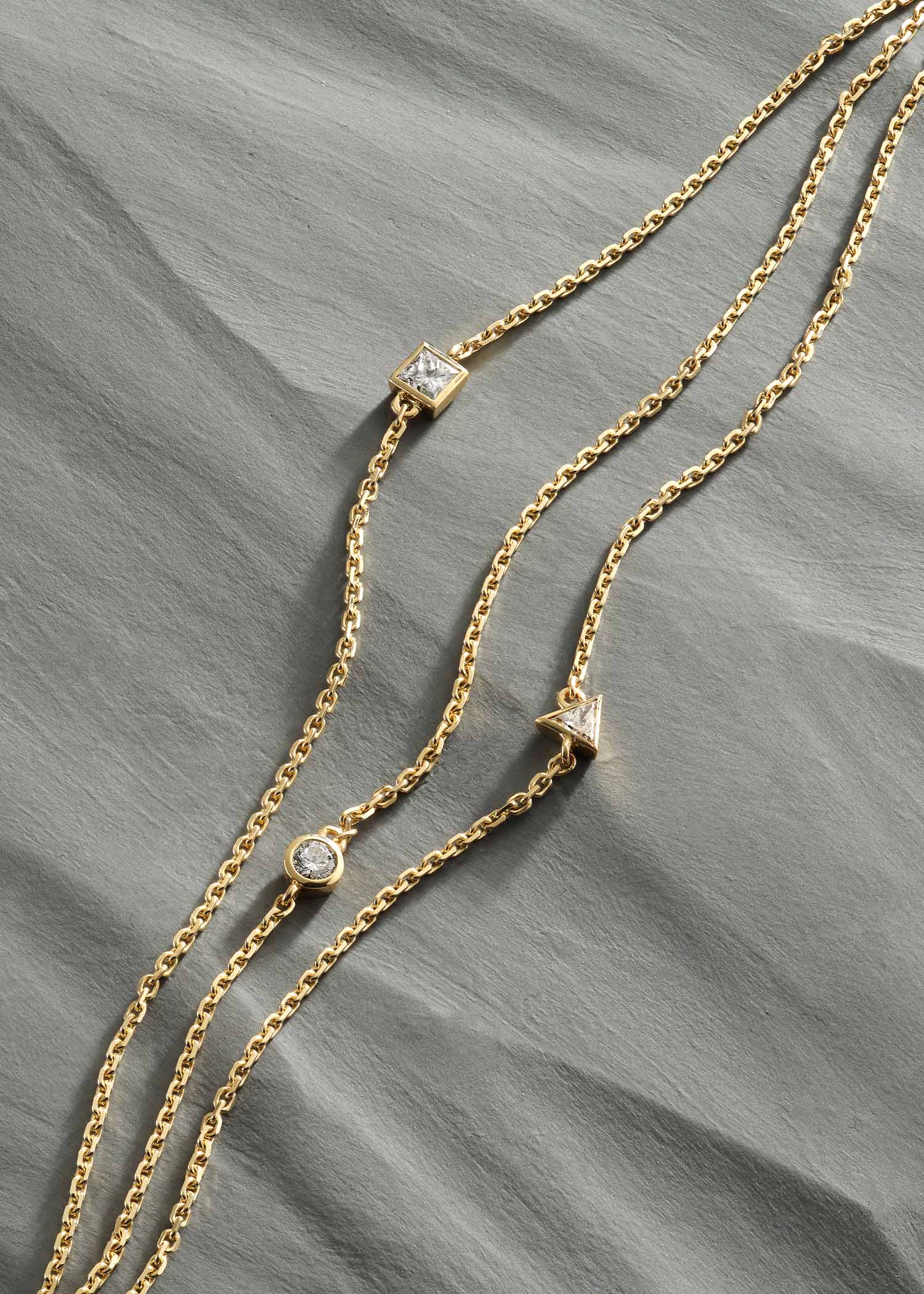 Delicate gold three-strand bracelet featuring round, triangle, and square-cut diamonds, photographed on a textured gray surface.