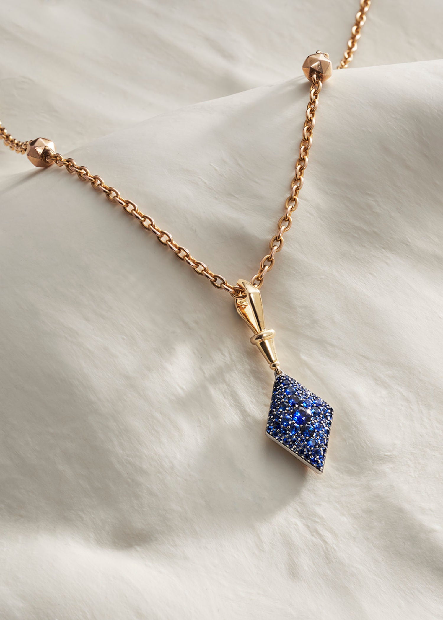 Three gold diamond-shaped pendants in green, pink, and blue gemstone settings displayed on a soft white fabric background.