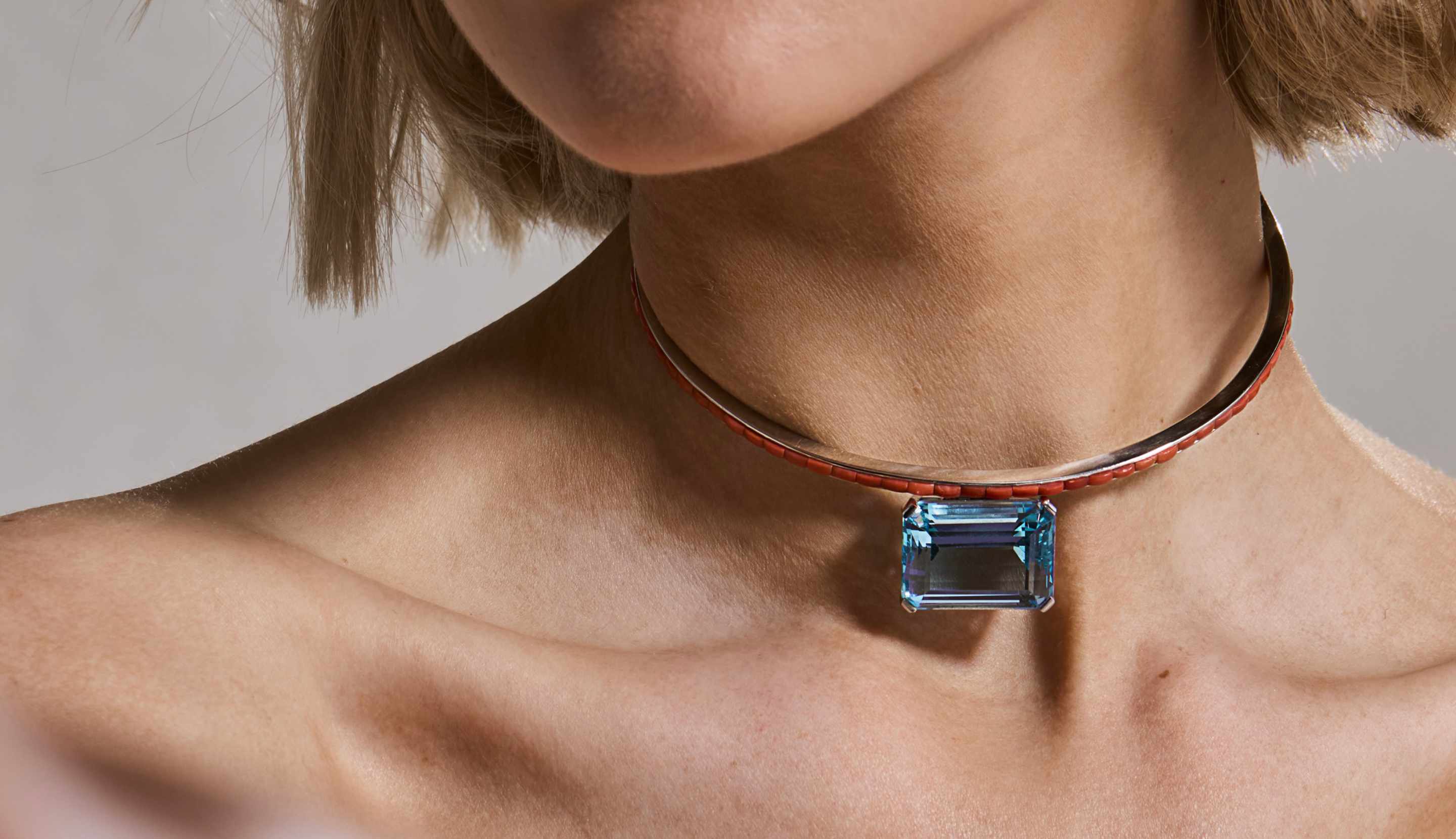 Blonde woman with slicked-back hair wearing a black strapless top and a choker necklace with a large light blue gemstone.