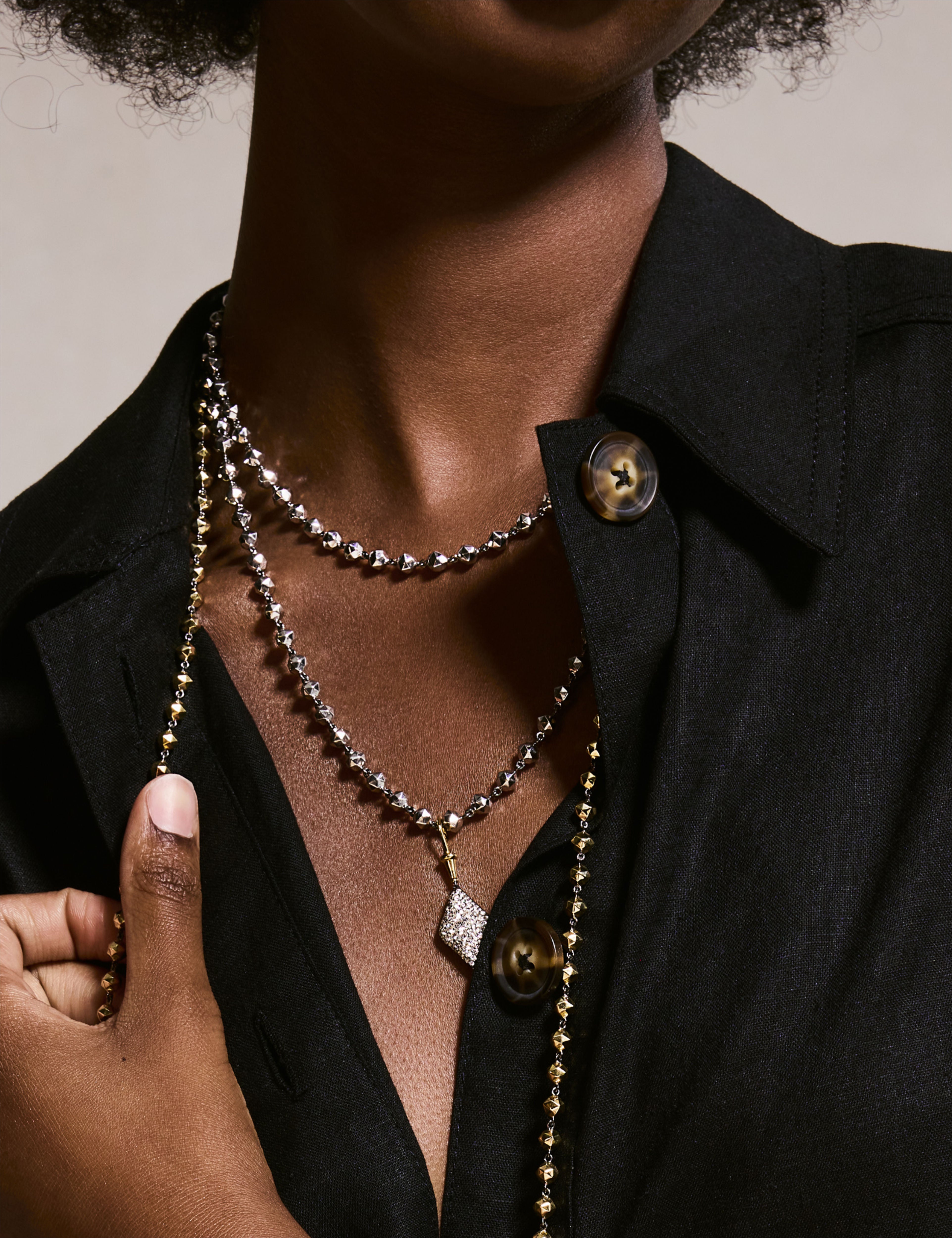 Model wearing layered gold bead necklaces with a diamond-shaped pendant and matching gold earrings, paired with a brown top.