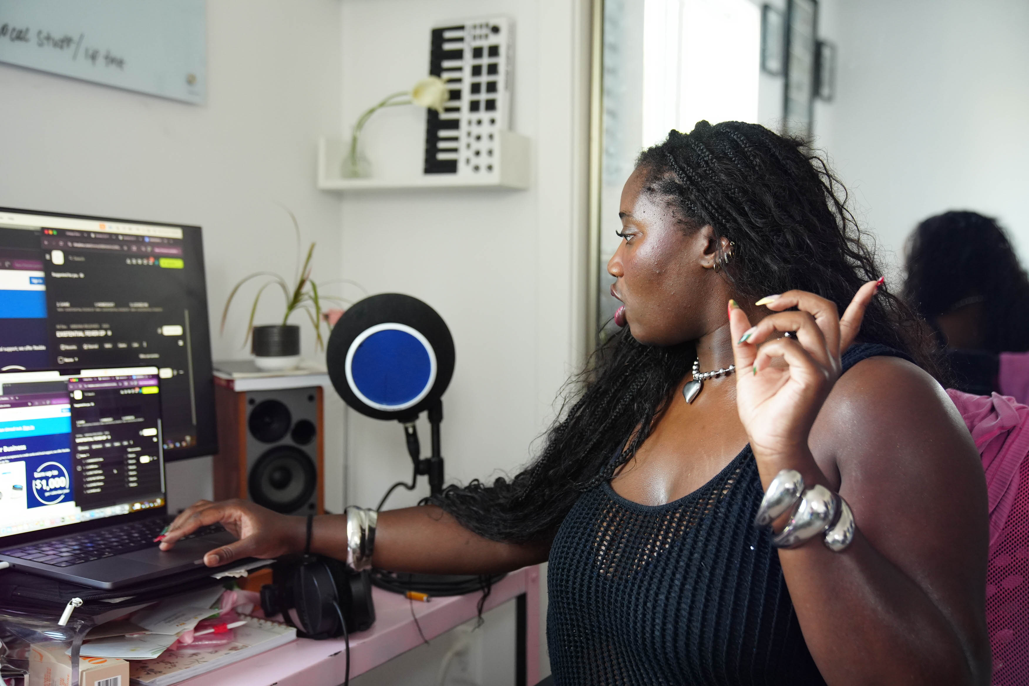A young woman is gesturing with one hand, the other hand on her laptop and she's surrounded by music equipment, sitting in a chair.