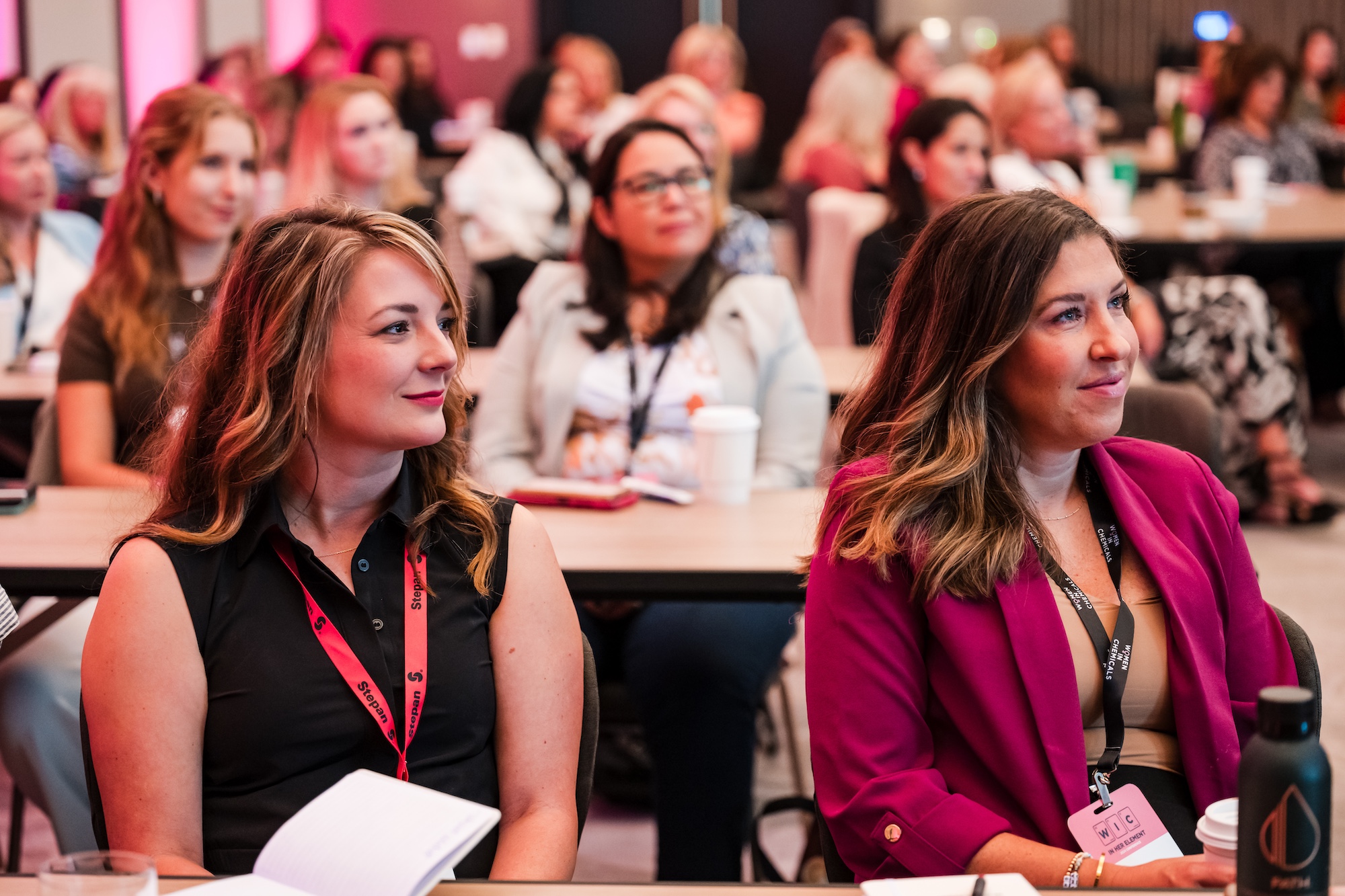 A group of women, in business casual attire, sit at a table with notebooks open, facing and listening to a speaker off-camera.