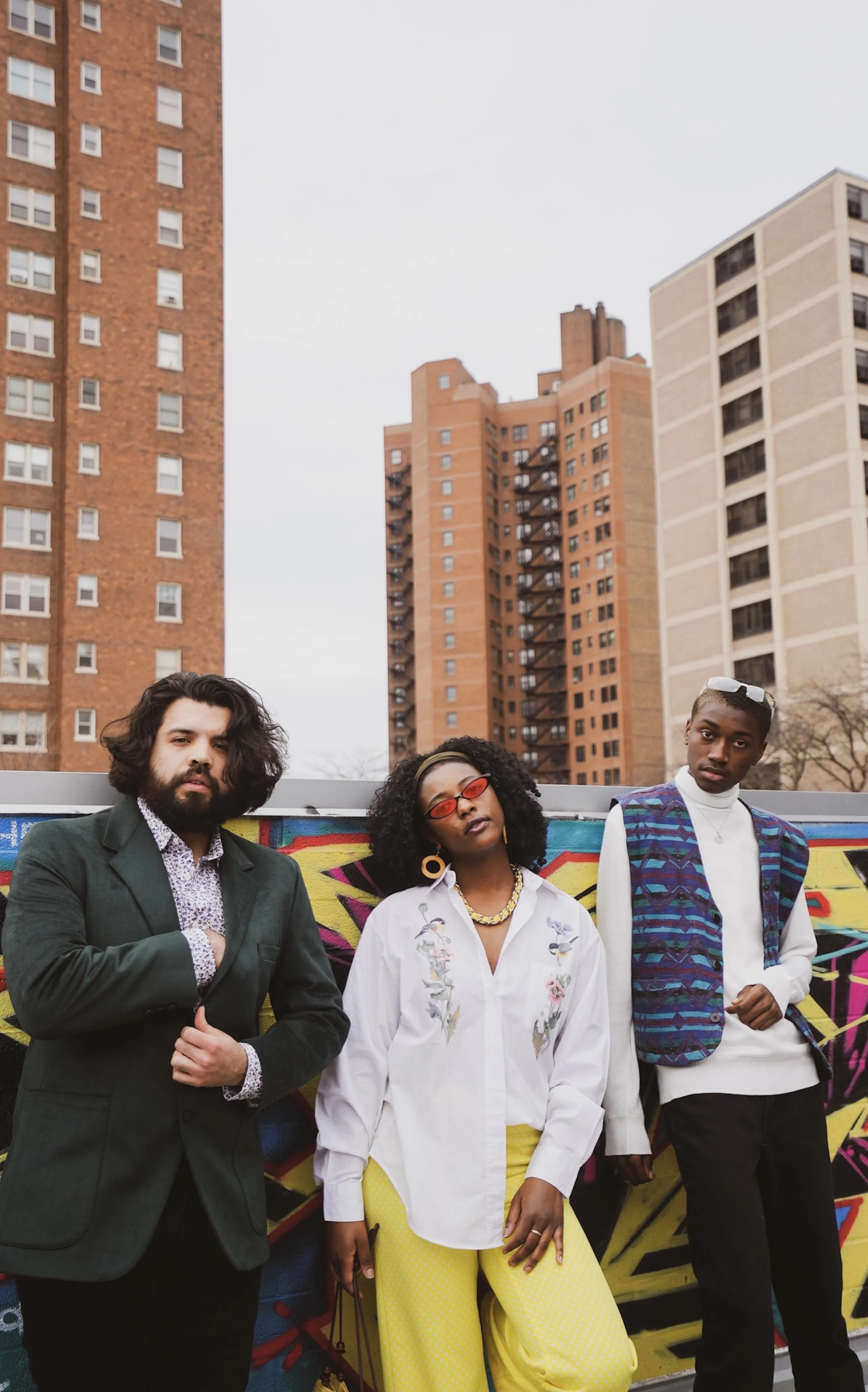 3 teenagers pose on a rooftop outside, tall apartment buildings in the background.