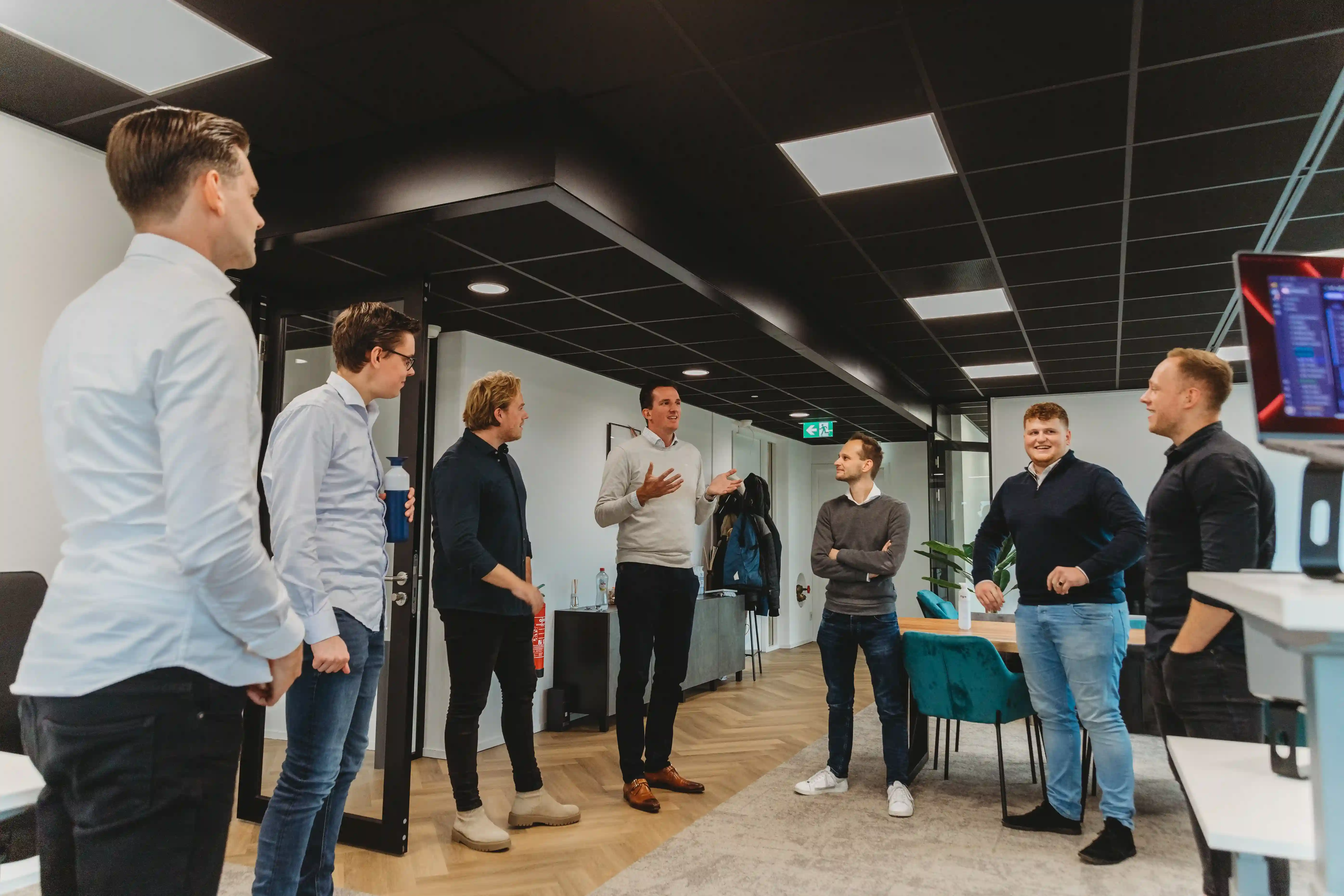 Seven men standing and conversing in a modern office space with wooden floors and black ceiling panels.