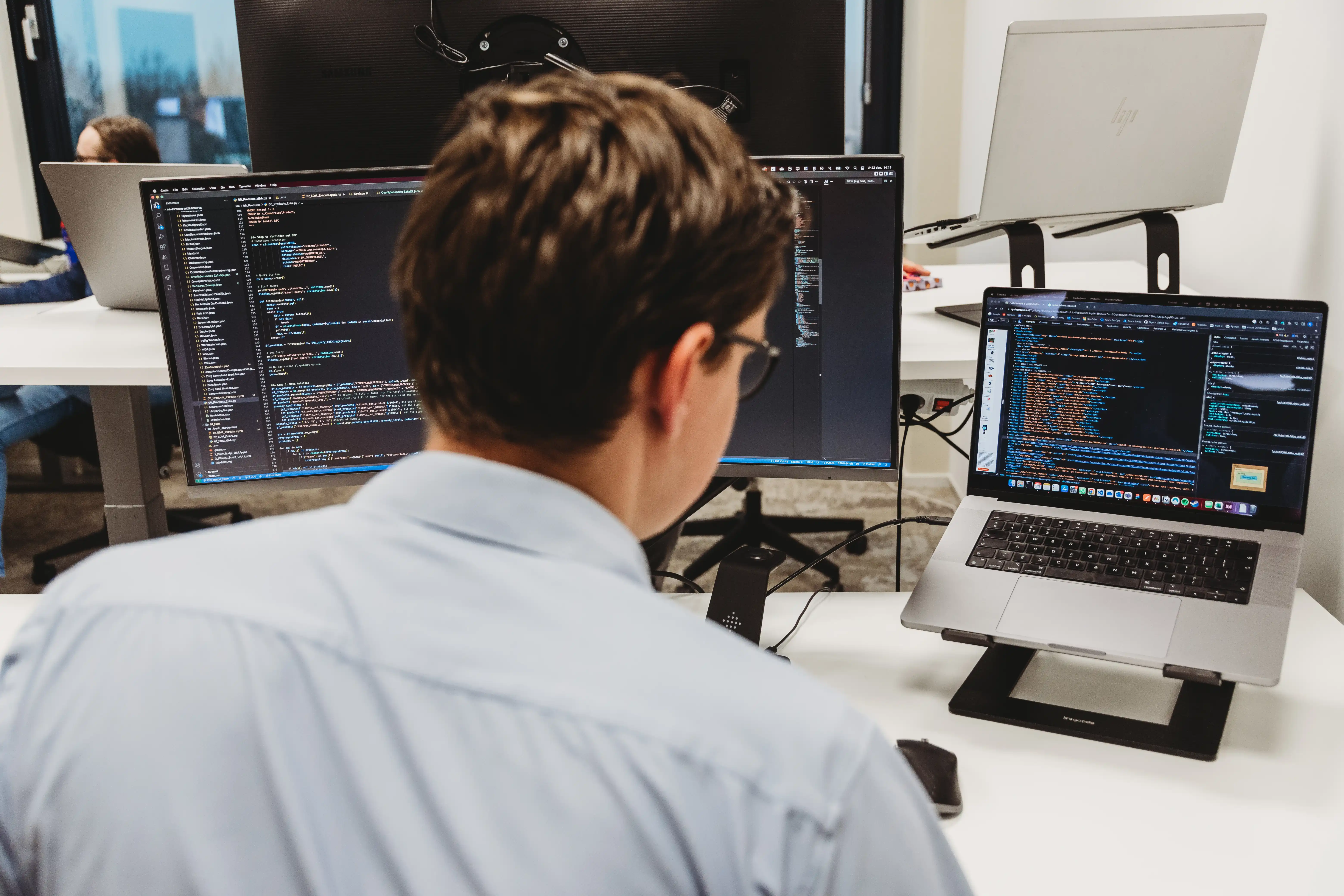 Man wearing glasses and a light blue shirt working on coding displayed on a laptop and a large monitor in an office.