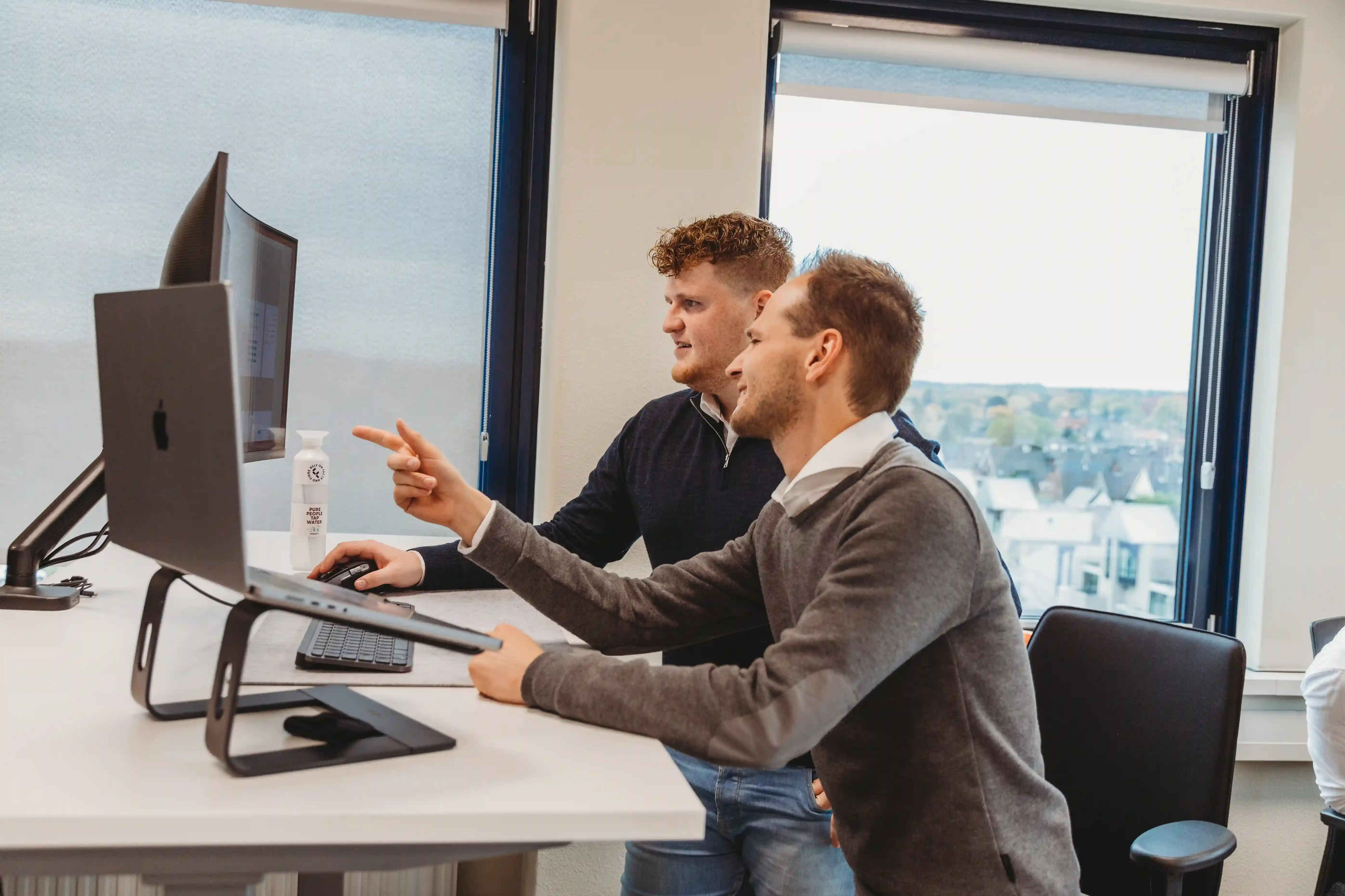 Two men collaborating and pointing at a computer screen in an office with large windows.