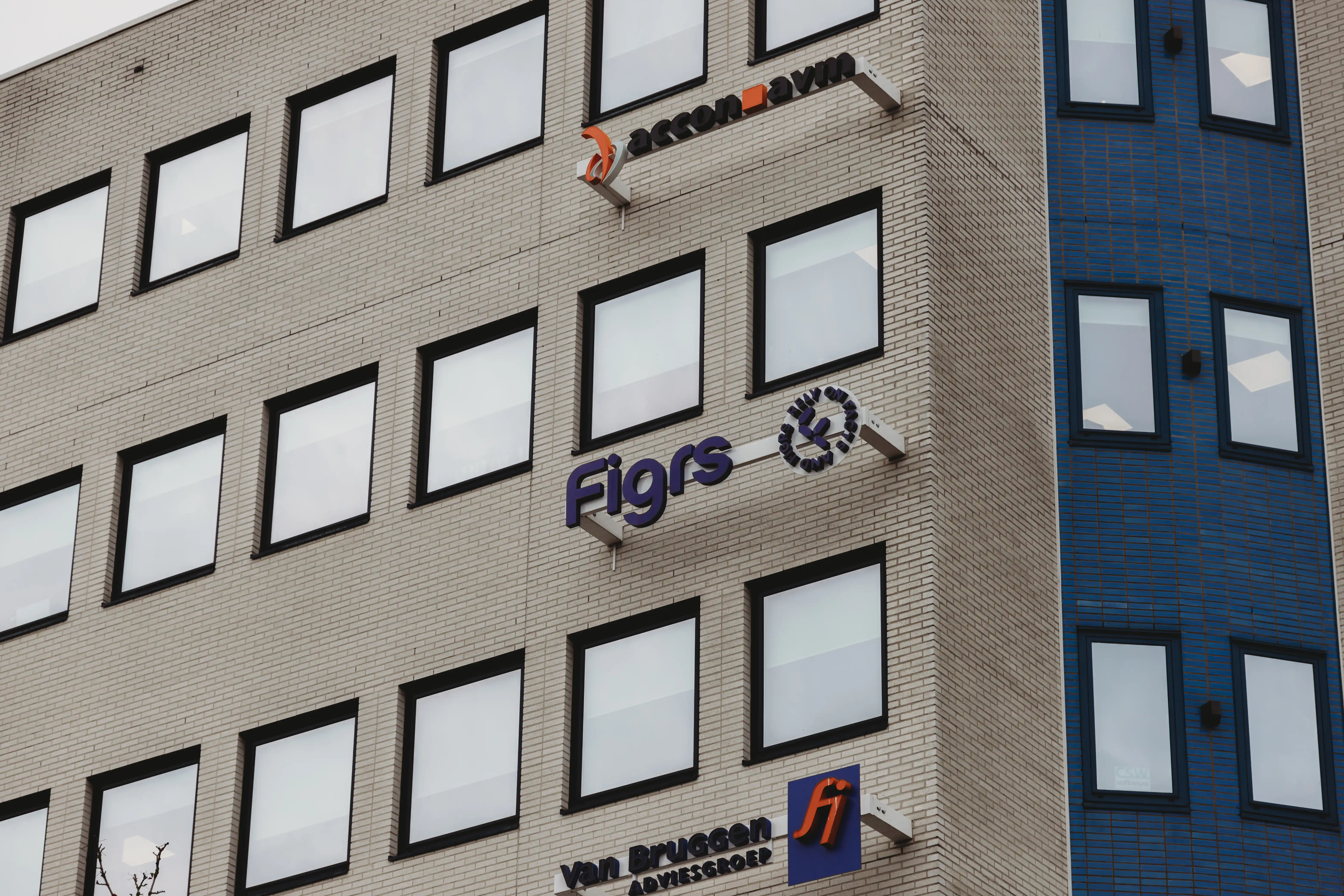 Exterior view of a brick office building corner with multiple square windows and business signs reading 'accon avm,' 'Figrs,' and 'Van Bruggen Adviesgroep.'