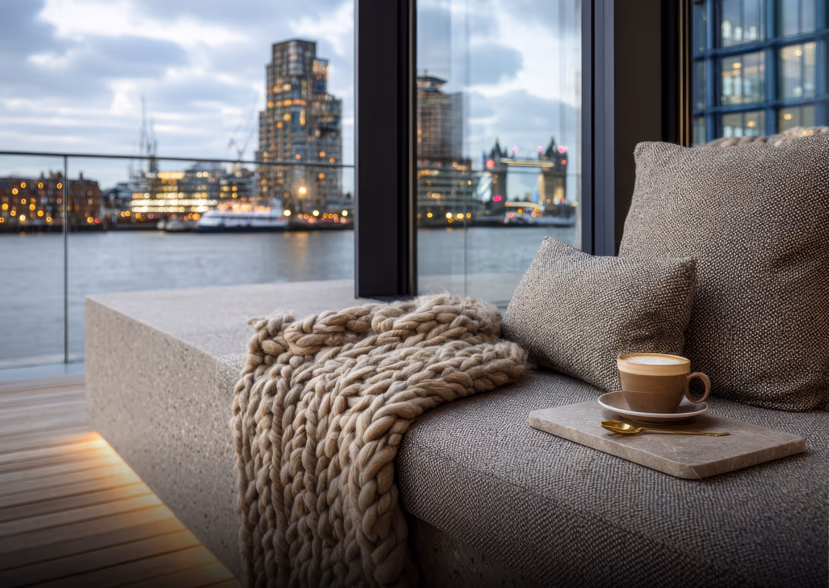 Cozy seating area with gray cushions and a thick beige knitted blanket by a window overlooking a river and city skyline at dusk.