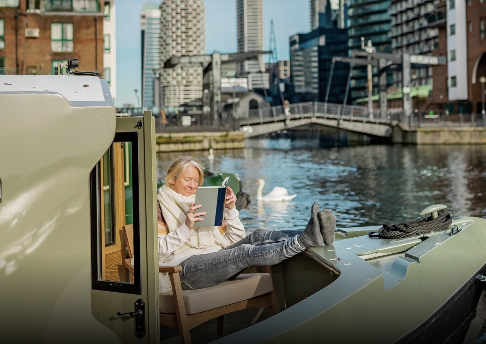 Woman in a warm scarf sitting on a chair with feet up on a boat, reading a book by the water with swans and city buildings in the background.