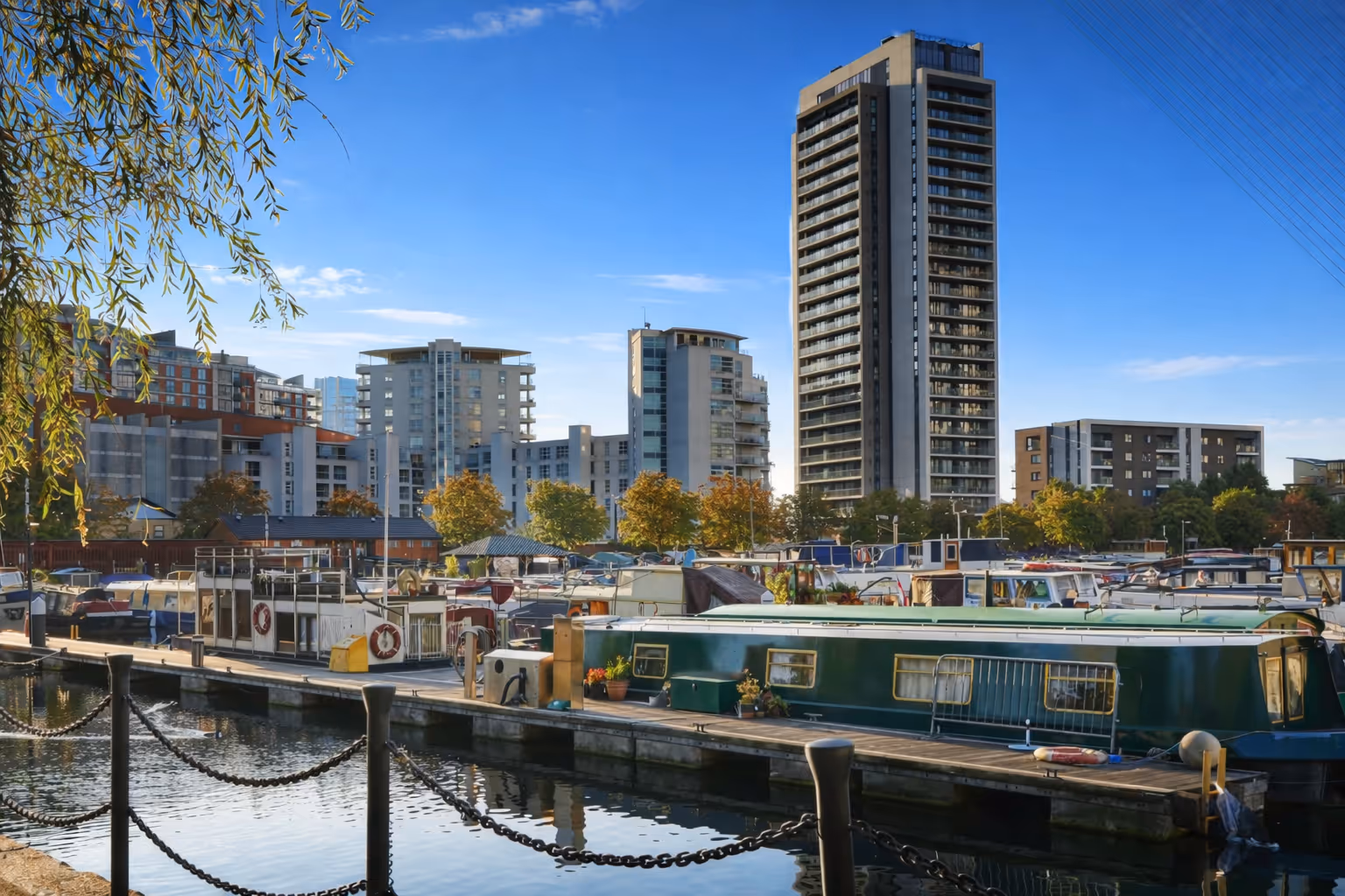 Marina with docked boats and narrowboats in calm water, with modern apartment buildings and a tall tower in the background.